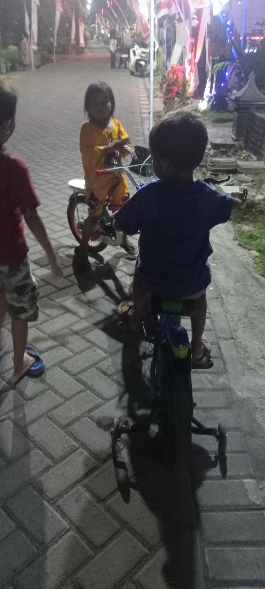 In Gempolklutuk, Indonesia, at dusk, three children are on a paved walkway. A boy in a dark blue t-shirt is riding a bicycle with training wheels, his back to the camera. To his left, a girl in a yellow shirt sits on a bicycle, looking forward. To the left of the girl, another child, mostly obscured by the girl, is visible from the chest down, wearing a red shirt and patterned shorts, and blue flip-flops. The walkway is made of interlocking gray bricks. In the background, there are decorated poles with red and white fabric, likely for a local celebration or event, and a white motorcycle is parked. The lighting suggests it is nighttime or late evening.