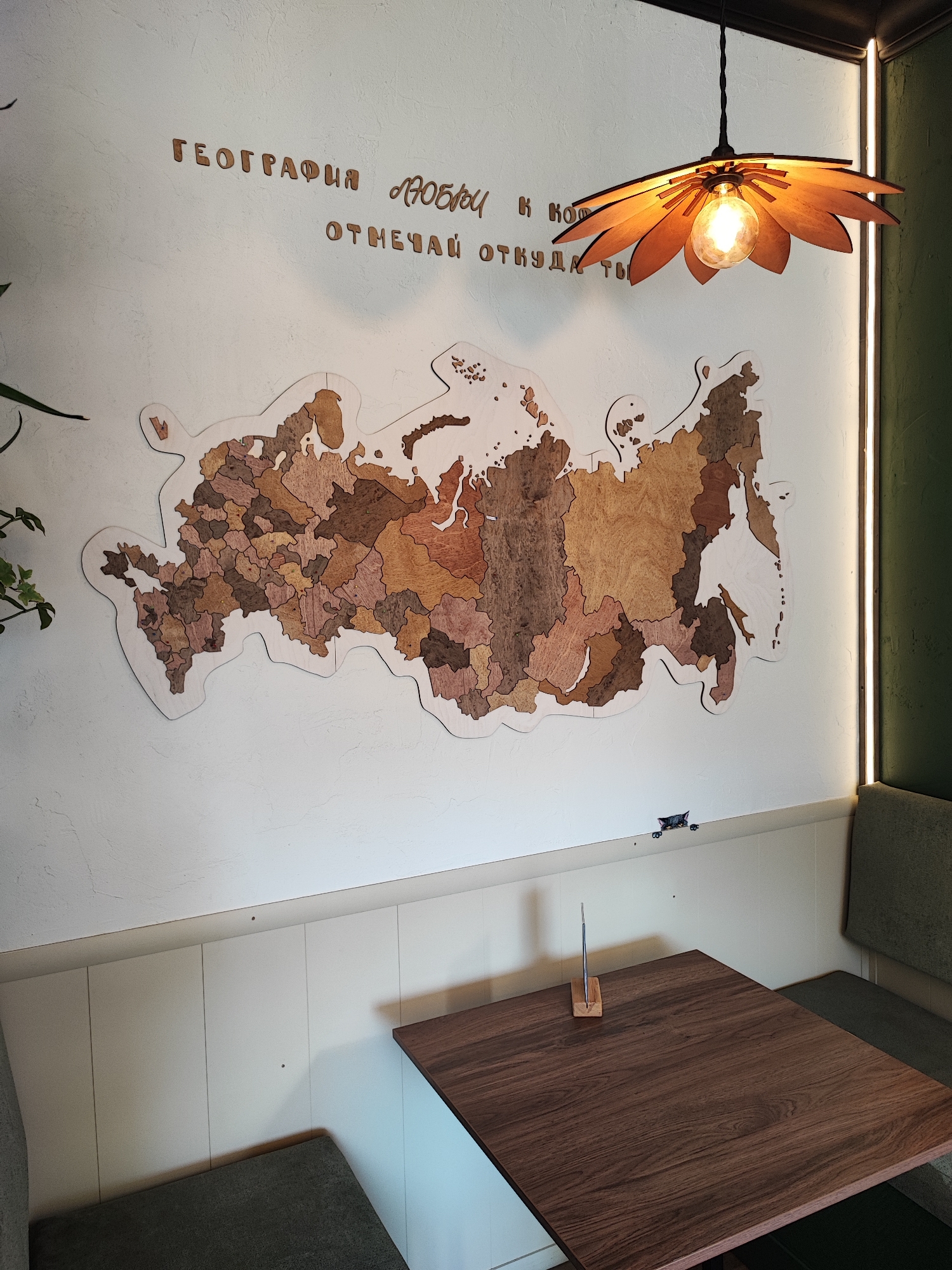 A wooden map of Russia, divided into regions, is mounted on a white textured wall. Above the map, wooden letters spell out a Russian phrase, "ГЕОГРАФИЯ ЛЮБОЧИ К КОТ ОТМЕЧАЙ ОТКУДА ТЫ" (Geography of Love to Kot, Mark from where you are). A decorative wooden pendant lamp with exposed bulbs hangs from the ceiling. Below the map, a dark wood table is set in a booth with green upholstered seating. The scene is illuminated by ambient lighting from the lamp and a vertical light strip on the right side of the wall. No people are visible in the image. The overall setting appears to be a cafe or restaurant interior.