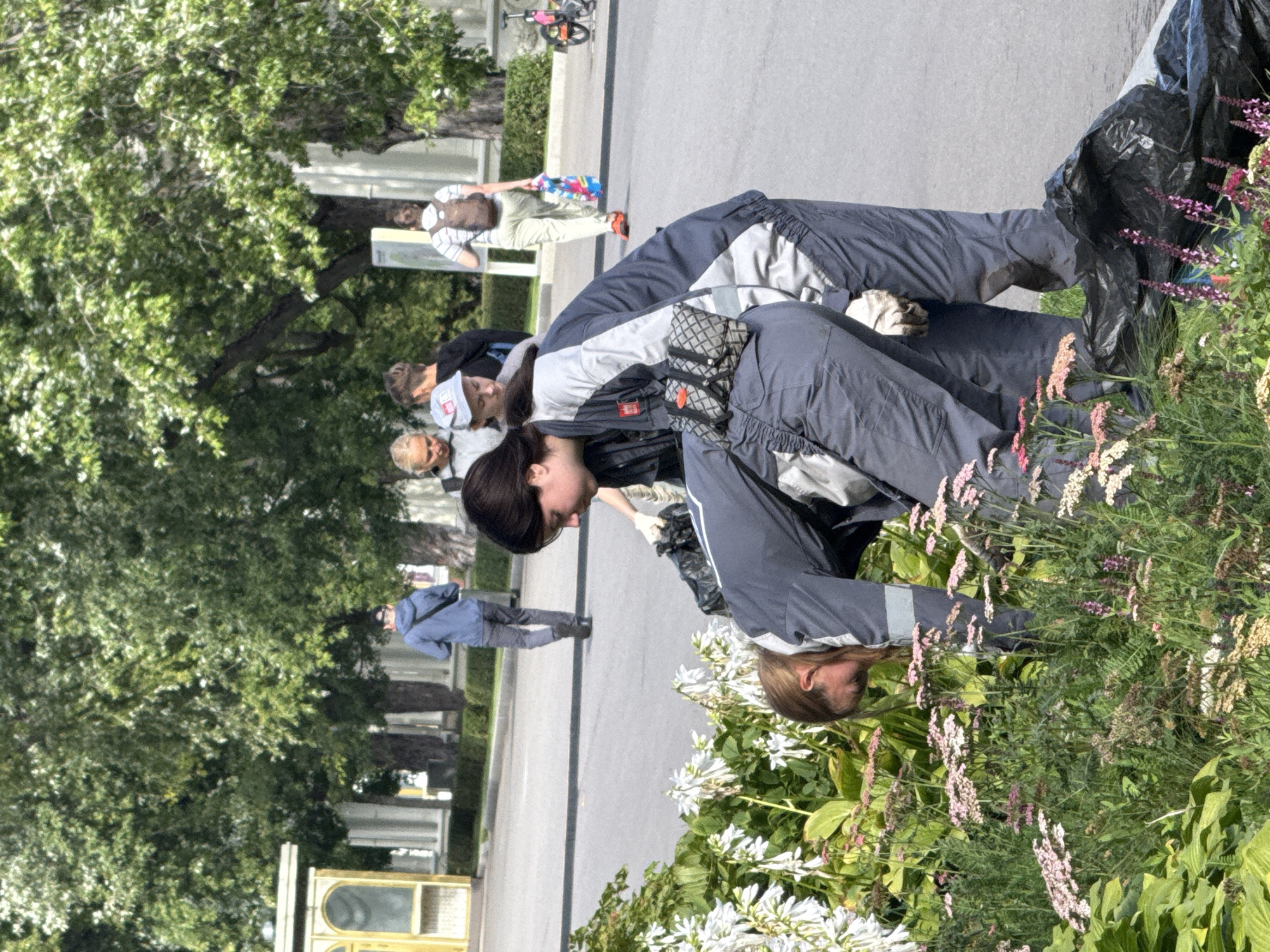 In an outdoor urban park, two young women are engaged in gardening activities. They are wearing matching grey work suits with blue and white accents. One woman, in the foreground, is bent over, tending to a flower bed filled with pink and white blossoms. The other woman stands behind her, also in the process of gardening. In the background, several other individuals are visible, some walking and others observing. The environment includes lush green trees, a paved walkway, and a building with classical architectural elements. The scene suggests a community effort or a work crew engaged in maintaining the park's greenery. The presence of black trash bags near the women indicates they may be clearing debris or waste. There are no immediate signs to identify the specific park within Moscow, Russia.
