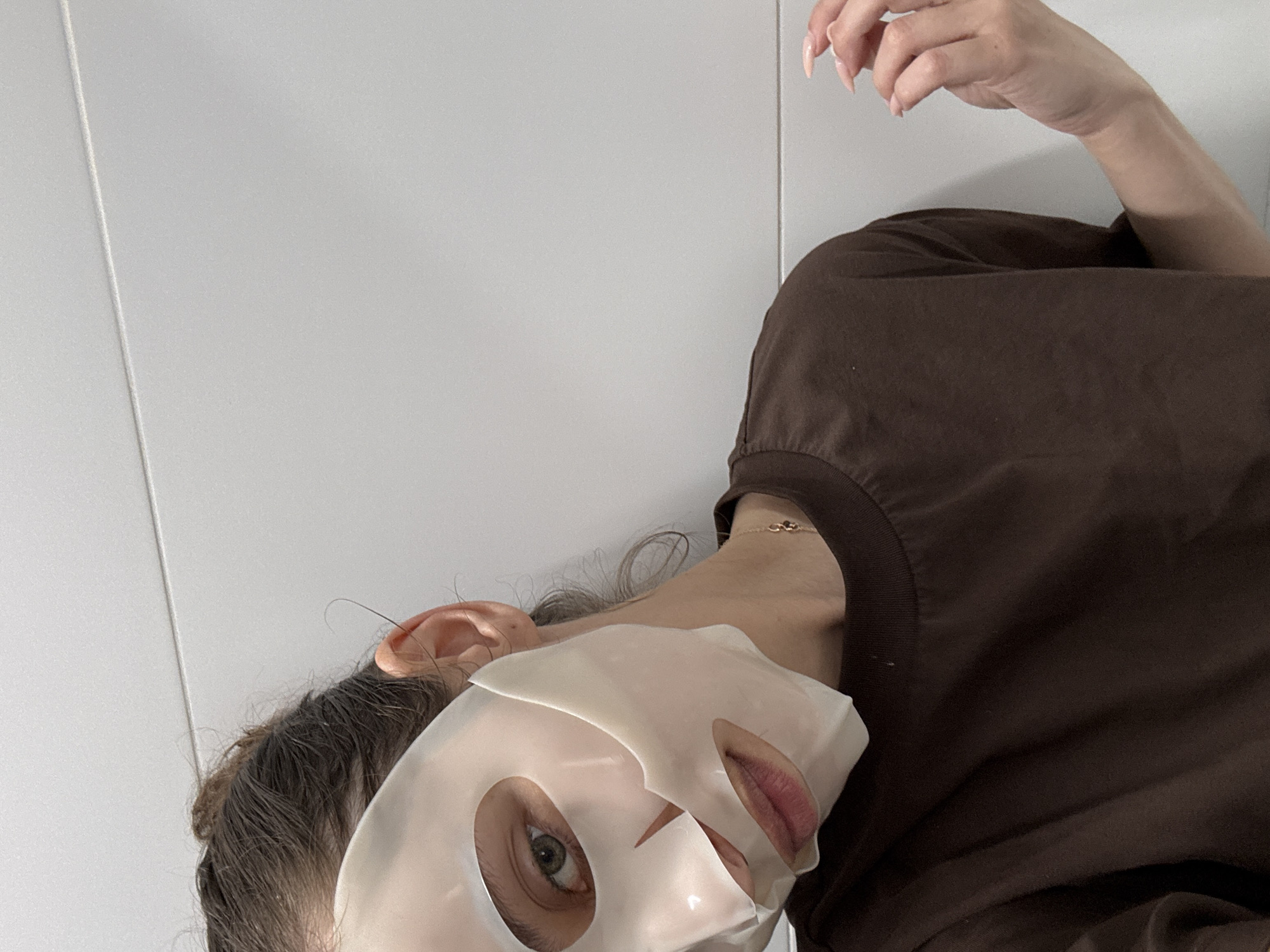 A woman wearing a sheet mask is photographed in front of a white wall. She is wearing a brown t-shirt and has a gold chain around her neck. Her right hand is raised near the top of the frame. The location appears to be indoors, but there are no elements to identify the city of Ramat Gan, Israel.
