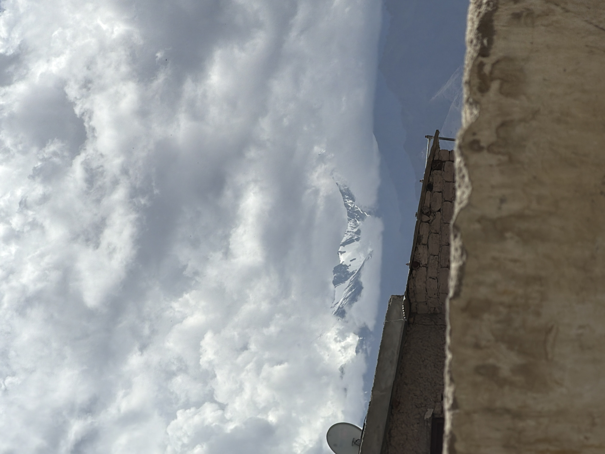 The image shows a cloudy sky over the city of Leh, India, with snow-capped mountains visible in the distance. In the foreground, there is a low wall made of rough stone and plaster. Behind the wall, a flat-roofed building is visible, constructed from stone and mud bricks, with a satellite dish mounted on its roof. The environment appears to be a rural or semi-urban area, with the mountains suggesting a high-altitude location. The lighting is diffused due to the cloud cover.