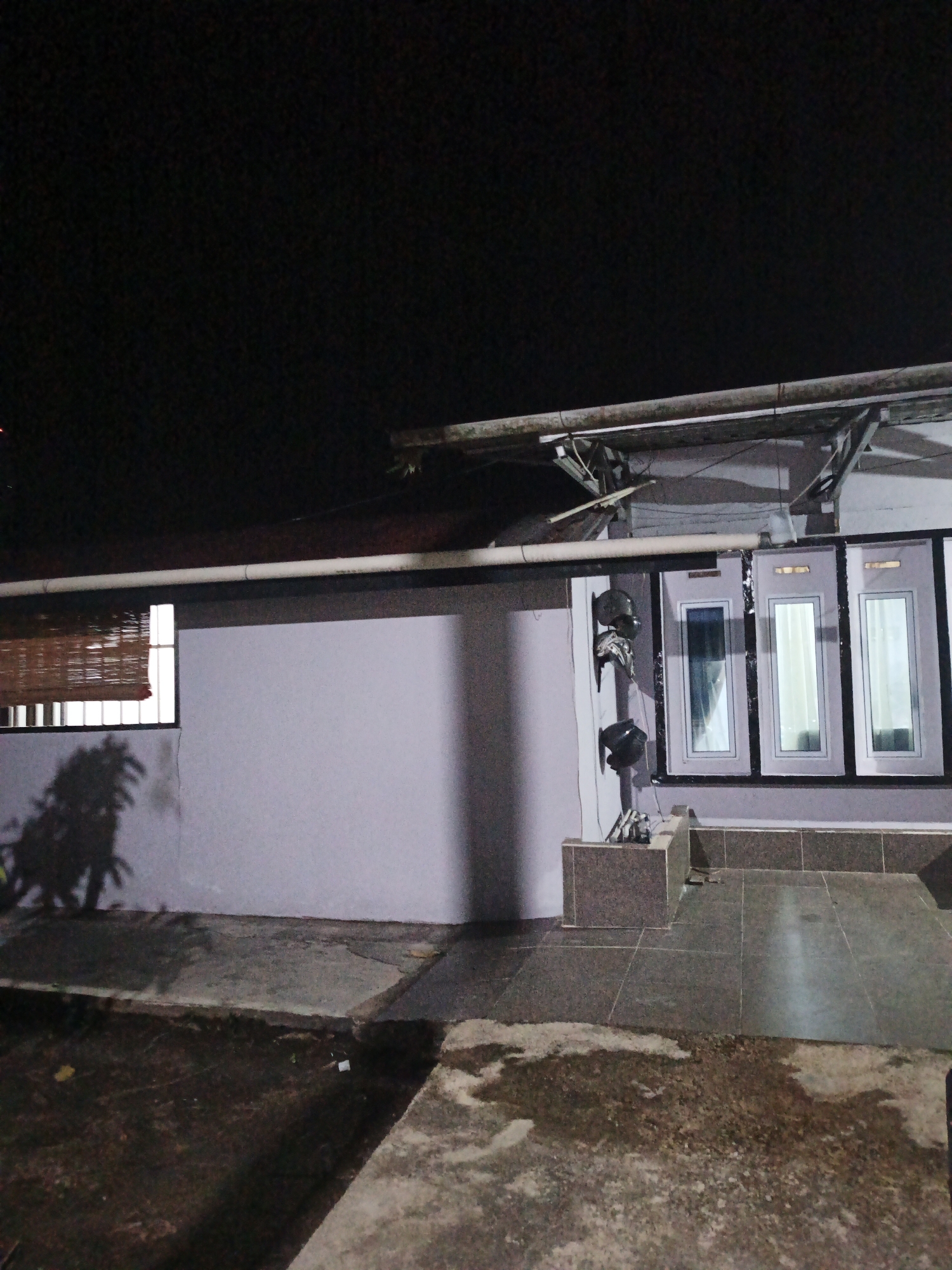 The image depicts the exterior of a building at night, with a dark sky overhead. A light-colored wall spans most of the frame, with a section of window covered by bamboo blinds on the left. To the right, there are three vertical windows with reflective glass. The building has a roof structure supported by metal beams. In front of the wall, there is a paved area with concrete and tiled sections, appearing somewhat wet or damp. A shadow of a tree is cast on the wall. Several dark objects, possibly helmets, are mounted on the wall to the left of the windows.