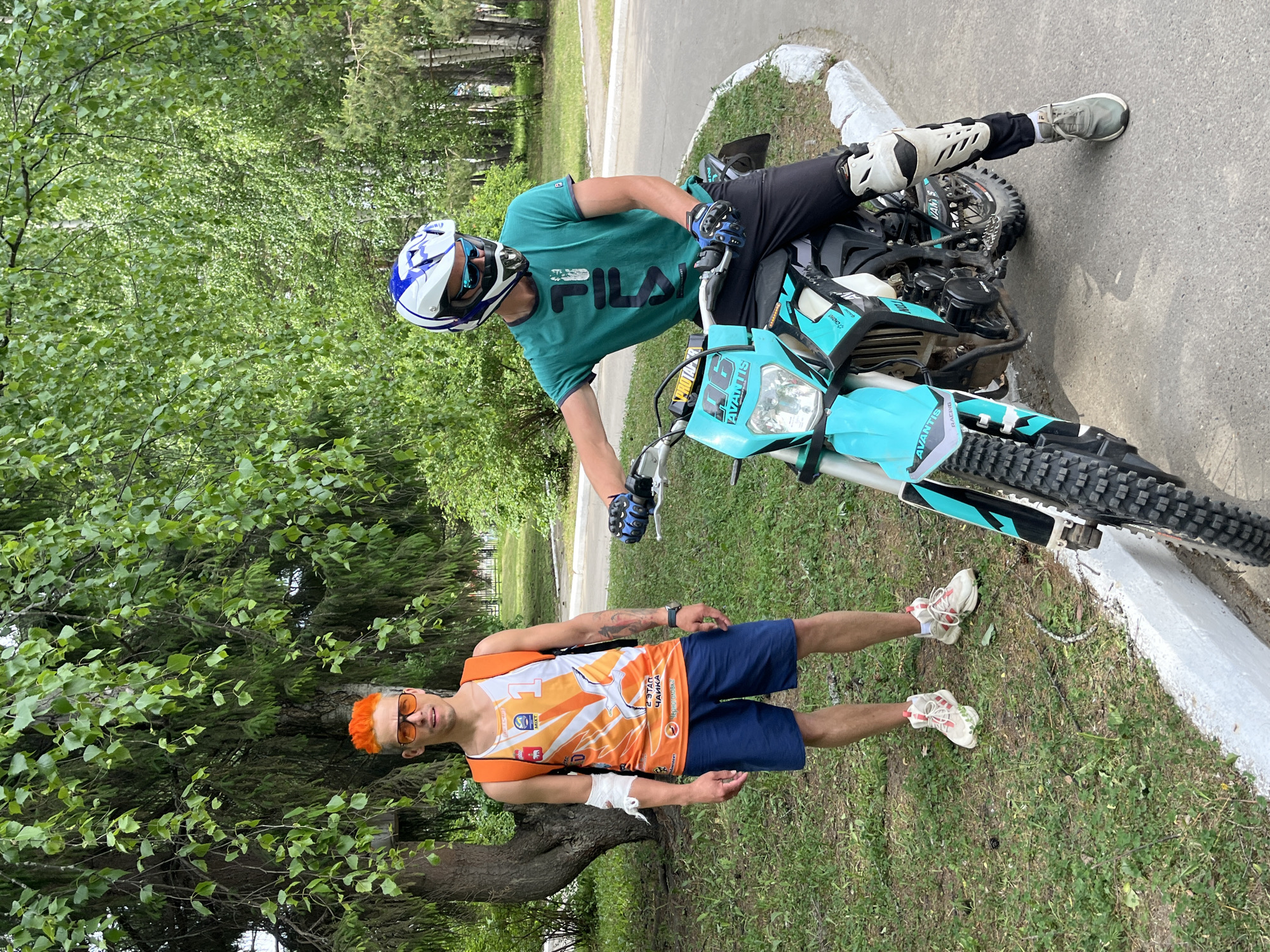 The image shows two men with a motorcycle outdoors. One man is sitting on a light blue and black motorcycle, wearing a helmet, a turquoise shirt with "FILA" printed on it, black pants, and protective gear on his knees. The other man stands beside a tree, wearing a white and orange tank top with Russian text and a bird graphic, blue shorts, orange sunglasses, and his hair is dyed orange. He has a bandage on his arm. The motorcycle has the text "AVANTIS RACING" on it and the number "06" on the front. The environment includes green grass, trees, and a paved road with a white curb. Text on the tank top indicates the location may be connected to the city of Chaikovsky, Russia.