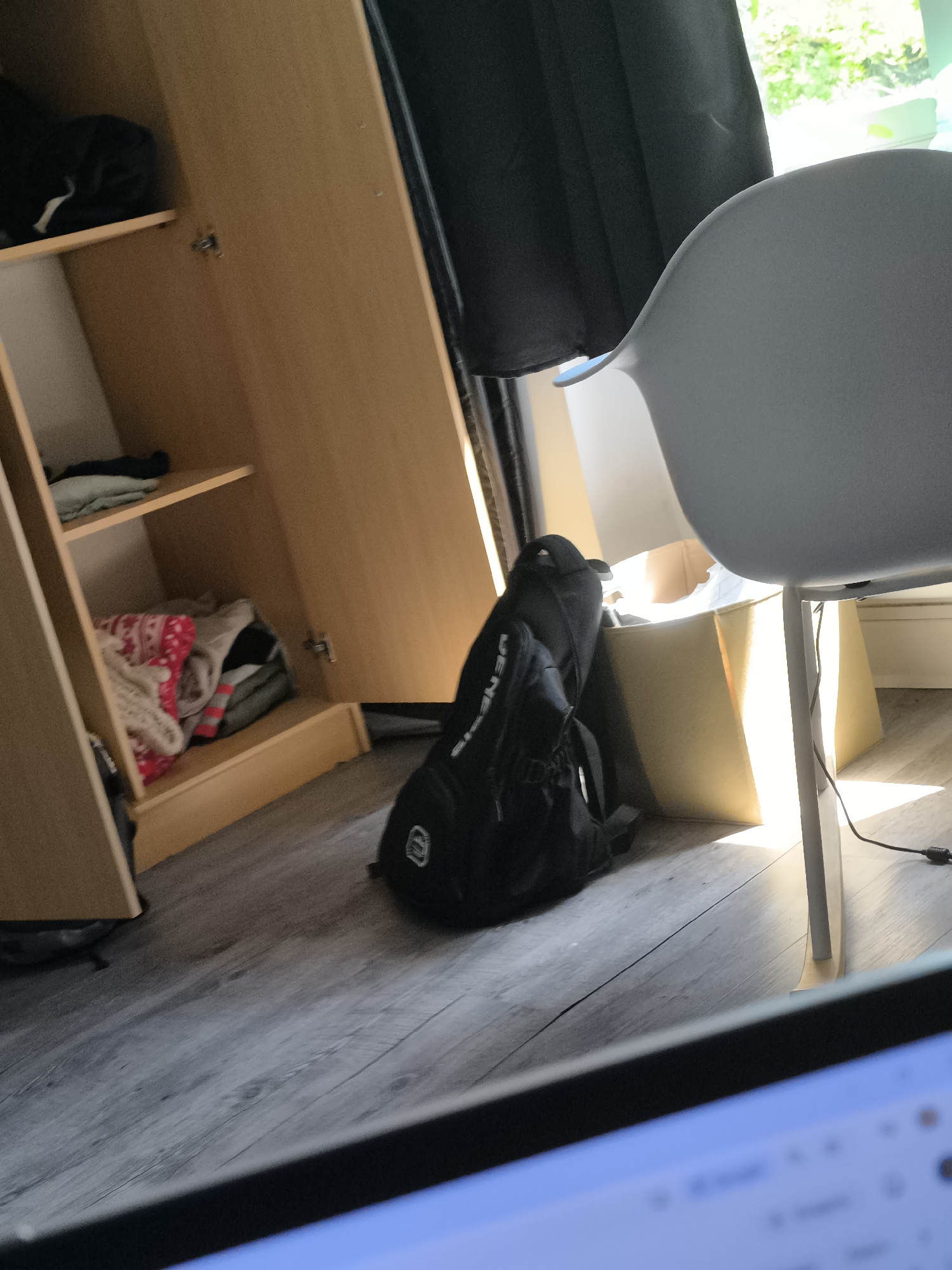 The image shows an interior scene with a partially open wooden wardrobe. Inside the wardrobe, clothing items are visible on shelves. Next to the wardrobe, a black backpack leans against a yellow cardboard box. A light gray chair is positioned near the box. The scene is illuminated by natural light coming from an unseen window. The image is partially obscured by a laptop screen in the foreground. The location cannot be determined to be Pélussin, France.