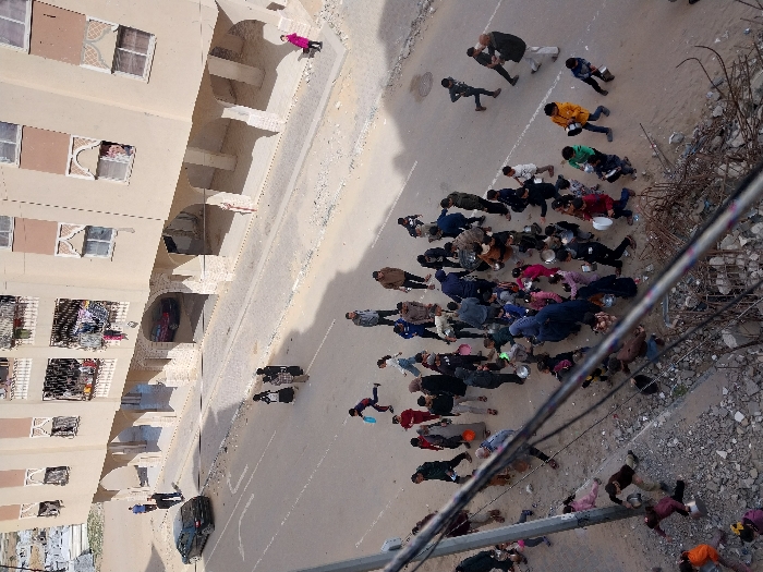 This image depicts an aerial view of a bustling street scene in Khan Yunis, Palestinian Territory, adjacent to a multi-story residential building. The building, rendered in a light beige tone, features multiple floors with balconies, some adorned with hanging laundry, and windows with decorative arch-like frames. Its ground floor incorporates a series of arched openings, one of which reveals a red car parked underneath.
The street, partially paved with asphalt and lined by a concrete sidewalk, is a focal point of activity. A large gathering of approximately fifty to sixty individuals, including numerous children and adults, occupies the street and the adjacent unpaved ground on the left. Many individuals in this crowd are holding empty bowls, pots, or plastic containers of various sizes, predominantly silver or white. Their collective movement appears directed, with some individuals running or walking briskly, suggesting a distribution or collection of essential items. Children are notably present throughout the crowd, many also carrying containers.
On the left side of the street, a black sedan is parked, with two men standing nearby. Further up the sidewalk on the right, a person in a pink coat stands alone. The immediate foreground shows a significant amount of debris, including rubble, dirt, and twisted rebar, indicating a rough or possibly damaged ground surface. Utility poles and wires partially obscure the lower left portion of the frame, indicating the elevated perspective of the photograph. The sky above is clear, suggesting daylight. In the distant background, open, arid terrain is visible.