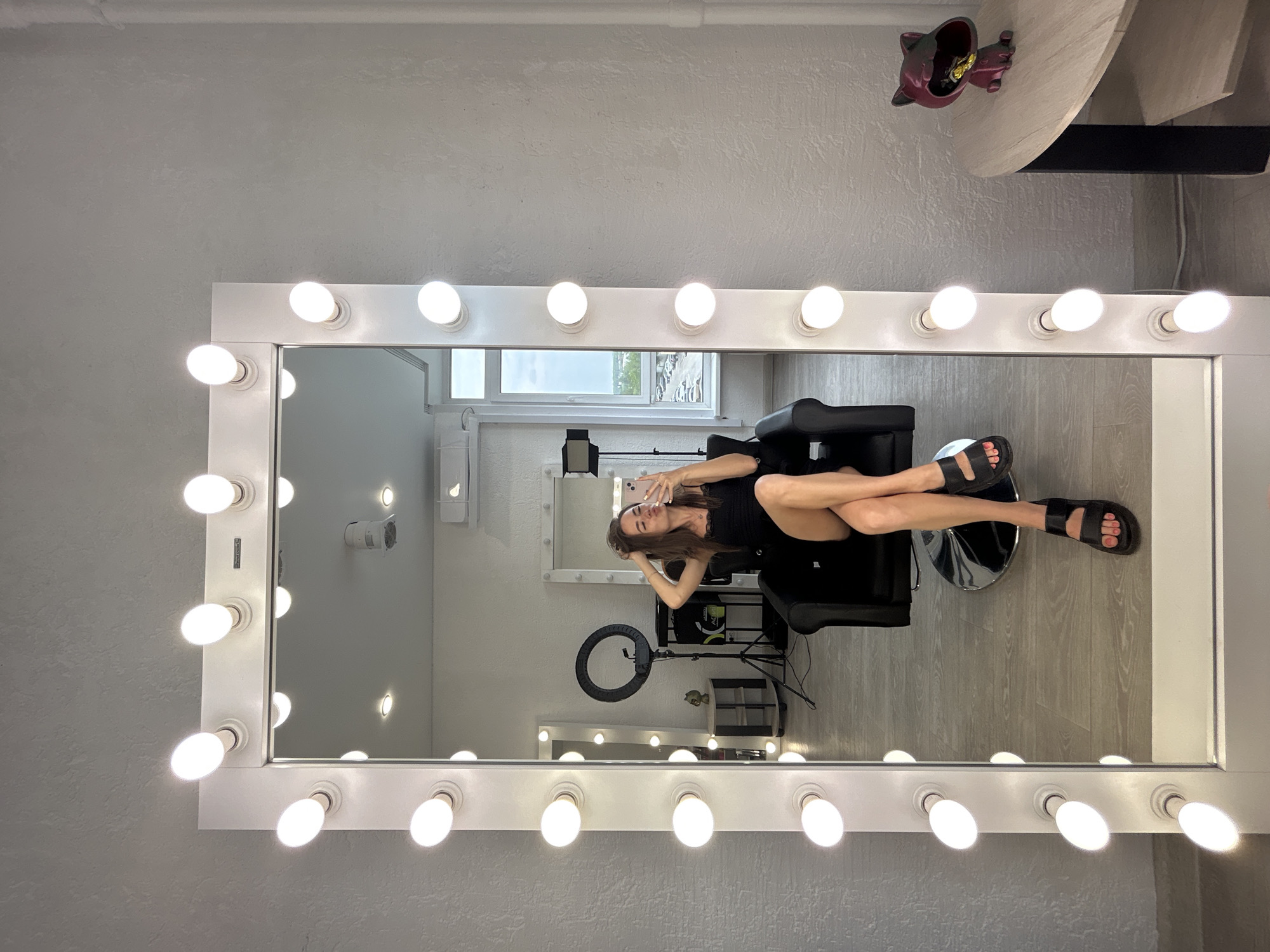 The image shows a woman sitting in a black chair in front of a large mirror with lights. She is wearing a black dress and black sandals. She is holding her phone and taking a selfie. In the reflection of the mirror, we can see a ring light, a small table with a cat-shaped decoration, and a window with a view of a parking lot and some buildings. The room appears to be a beauty salon or a dressing room.