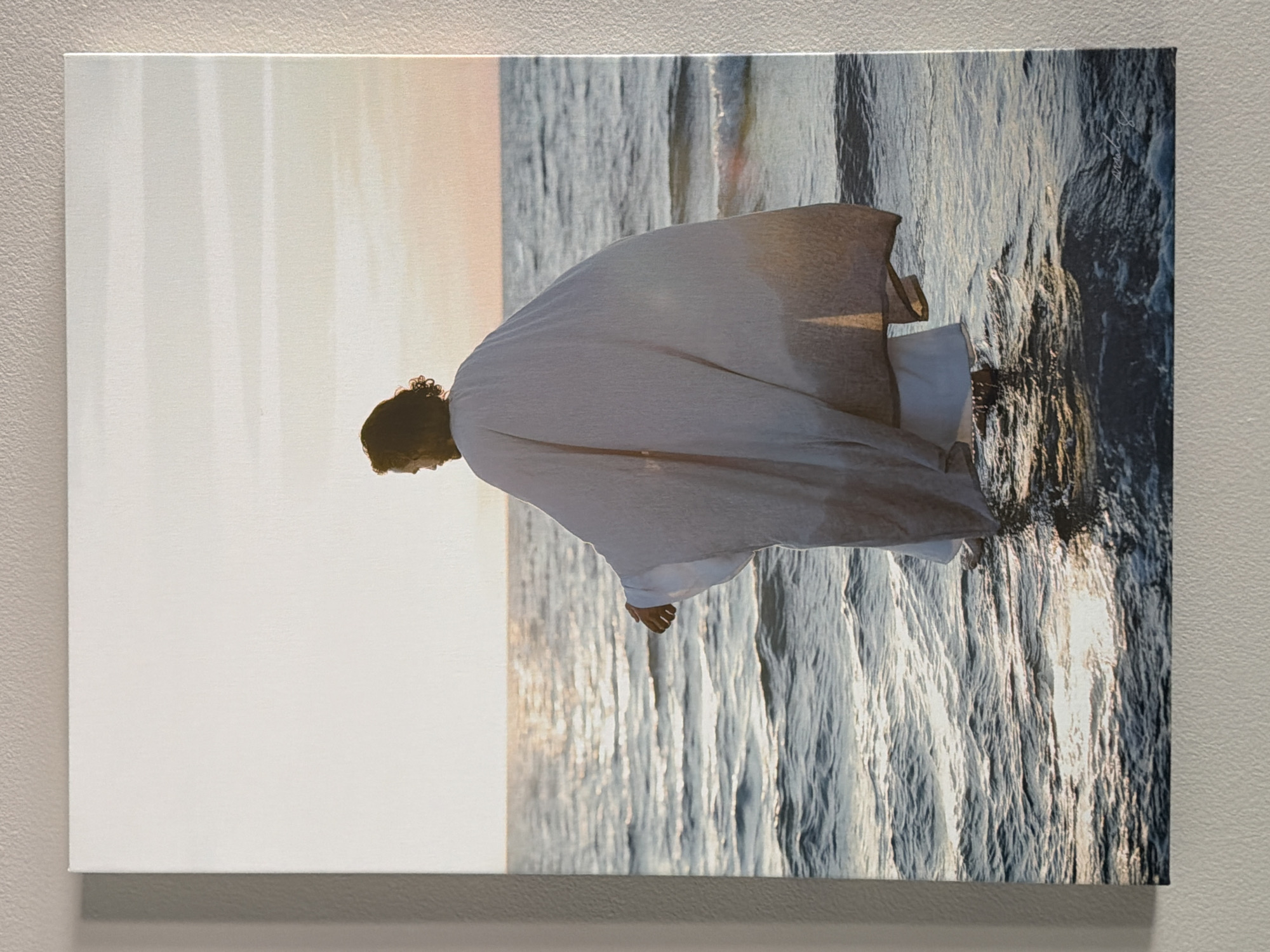 A canvas print depicts a person from behind, standing in the ocean during sunset. The individual is wearing a long, flowing white robe with a gradient effect, darkening towards the hem. Their dark hair is visible, and their arms are outstretched slightly. The sun, low on the horizon, casts a warm glow across the sky and reflects brightly on the rippling water. Waves gently lap around the person's legs, which are partially submerged. The scene suggests a serene and contemplative moment. The canvas is displayed on a textured, neutral-colored wall, possibly in a home in Wellford, United States of America.