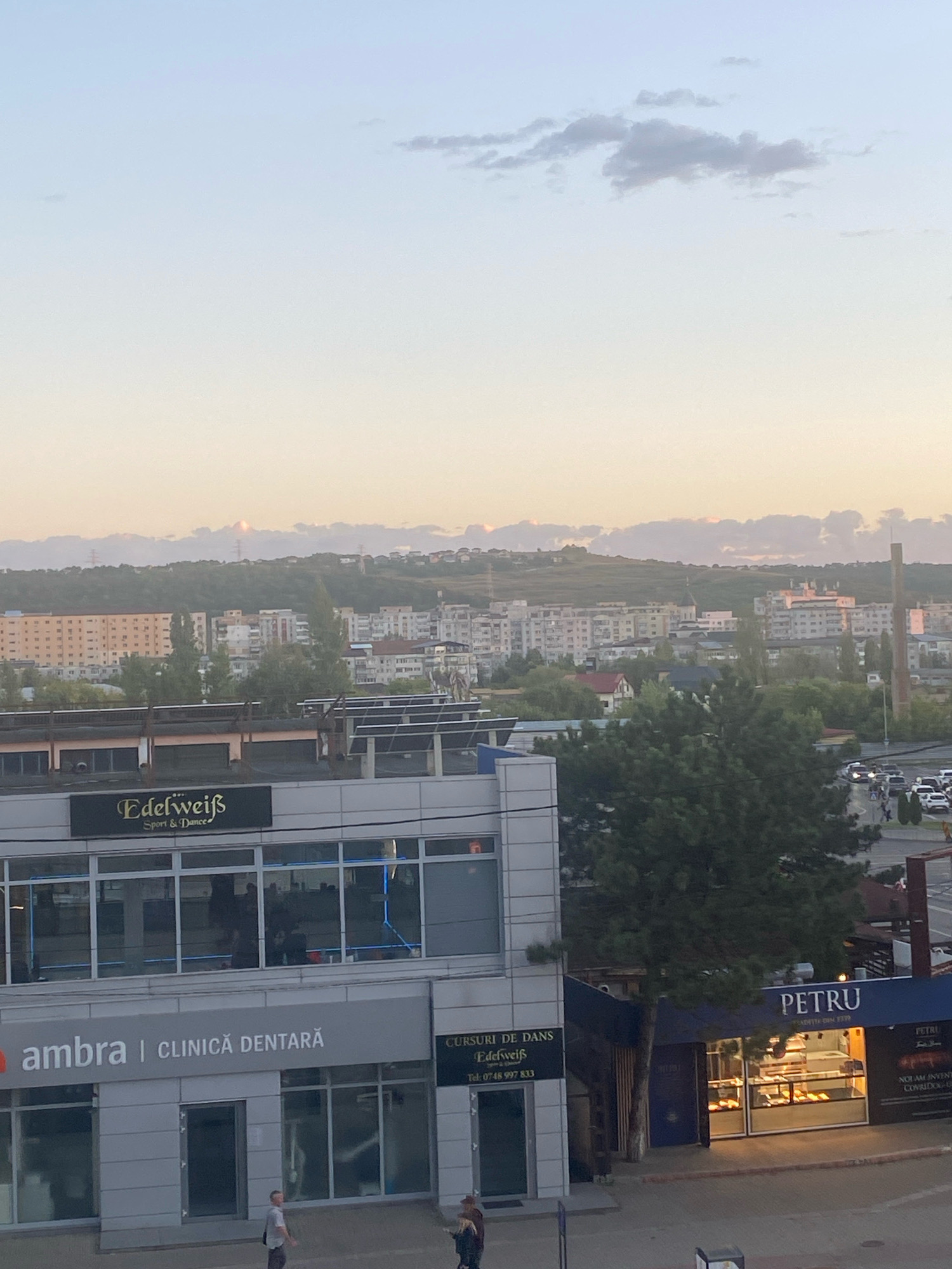 The image depicts a cityscape at sunset, likely in Iași, Romania, given the signage. In the foreground, there's a modern building with several businesses. One sign reads "Edelweiß Sport & Dance," another says "ambra | CLINICĂ DENTARĂ," and a third, partially visible, displays "CURSURI DE DANS Edelweiss" with a phone number. To the right, a bakery with the name "PETRU" is illuminated, displaying pastries in its window. Behind these businesses, a larger residential area with multiple apartment buildings stretches towards a tree-covered hill. The sky is a soft gradient of blue and orange, with a few scattered clouds. A tall, slender chimney or industrial structure is visible in the middle ground. Several cars are parked and driving on a street in the mid-right of the image. Two individuals are walking on the sidewalk in the lower portion of the frame. The overall scene suggests an urban environment transitioning into evening.