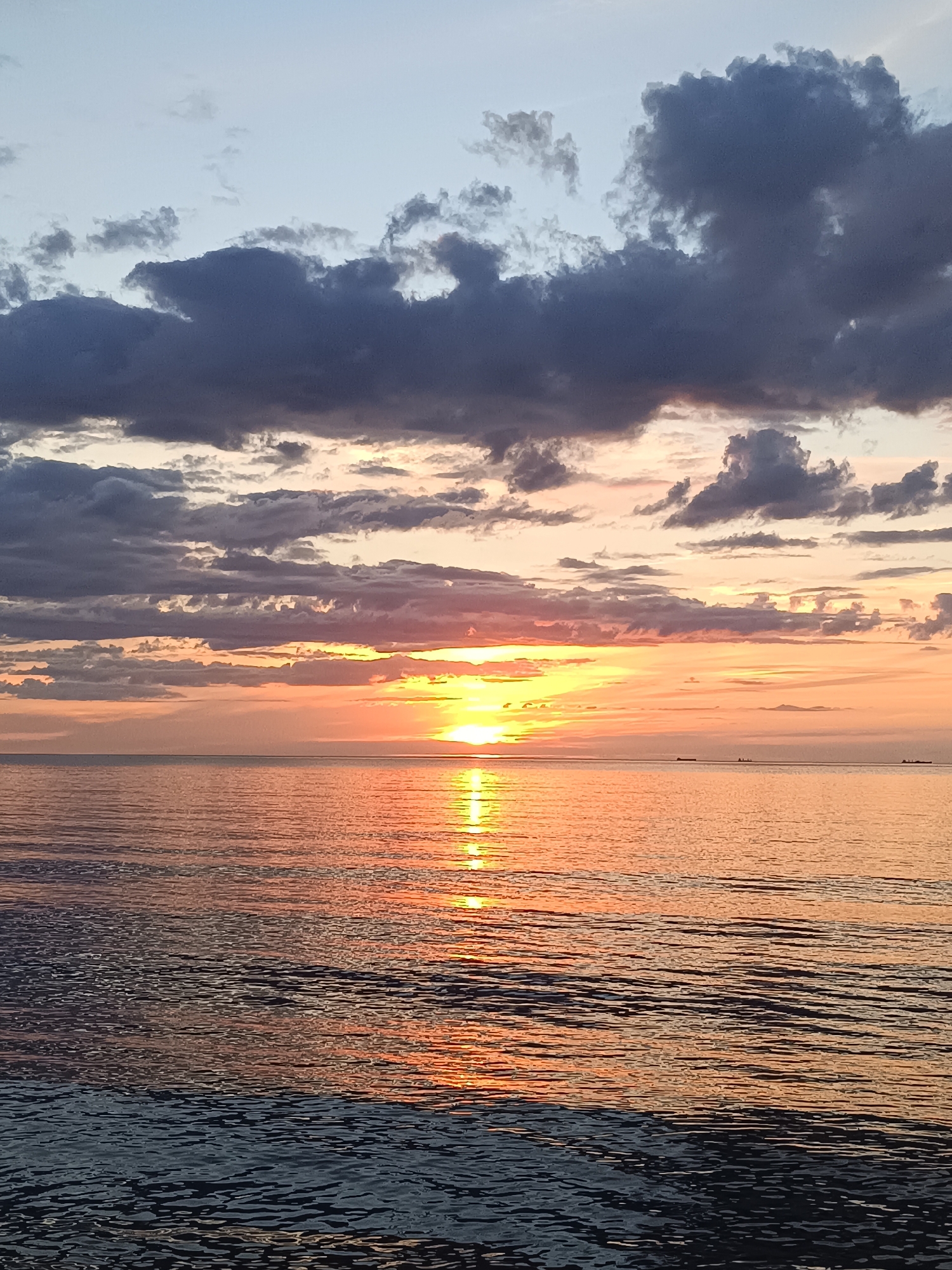 The image depicts a serene sunset over a calm body of water. The sun is low on the horizon, casting a warm, orange glow across the sky and reflecting on the water's surface. Dark, dramatic clouds are scattered across the upper portion of the sky, contrasting with the softer hues of the sunset. The water is rippled, creating a textured pattern of light and shadow. In the distance, several small, dark shapes that appear to be boats or ships are visible on the horizon. The overall scene is one of natural beauty and tranquility.