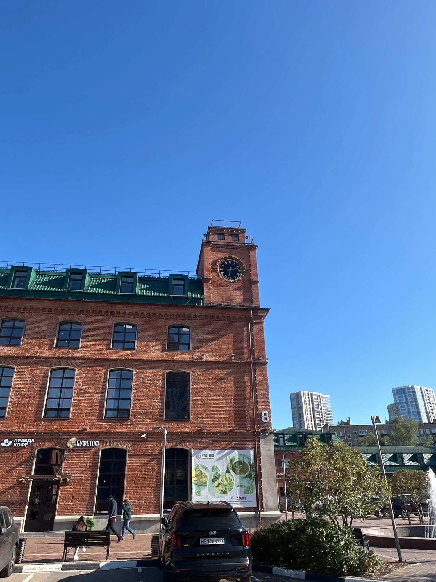 This is a vertical, eye-level, outdoor, daylight photograph of a red brick building with a green roof, featuring a prominent clock tower on the right side of the building. The building is in an urban environment, with other buildings visible in the background, including tall, modern apartment buildings. The sky is a clear, bright blue.
In the foreground, there is a paved area with parked cars, including a black Kia Sorento. There is a bench with two people sitting on it and two people walking past. A fountain is visible on the right side of the image. The building has multiple windows and signage, including logos for "ПРАВДА КОФЕ" and "БУФЕТОФ". There is also a poster with images of food.
Based on the signage, this location appears to be in Moscow, Russia, specifically at the address "Novodmitrovskaya Street, 5". The building itself is a former industrial building that has been repurposed for commercial use, housing cafes and other businesses. The architectural style suggests it may be a converted factory or warehouse. The presence of modern apartment buildings in the background indicates a developing urban area.