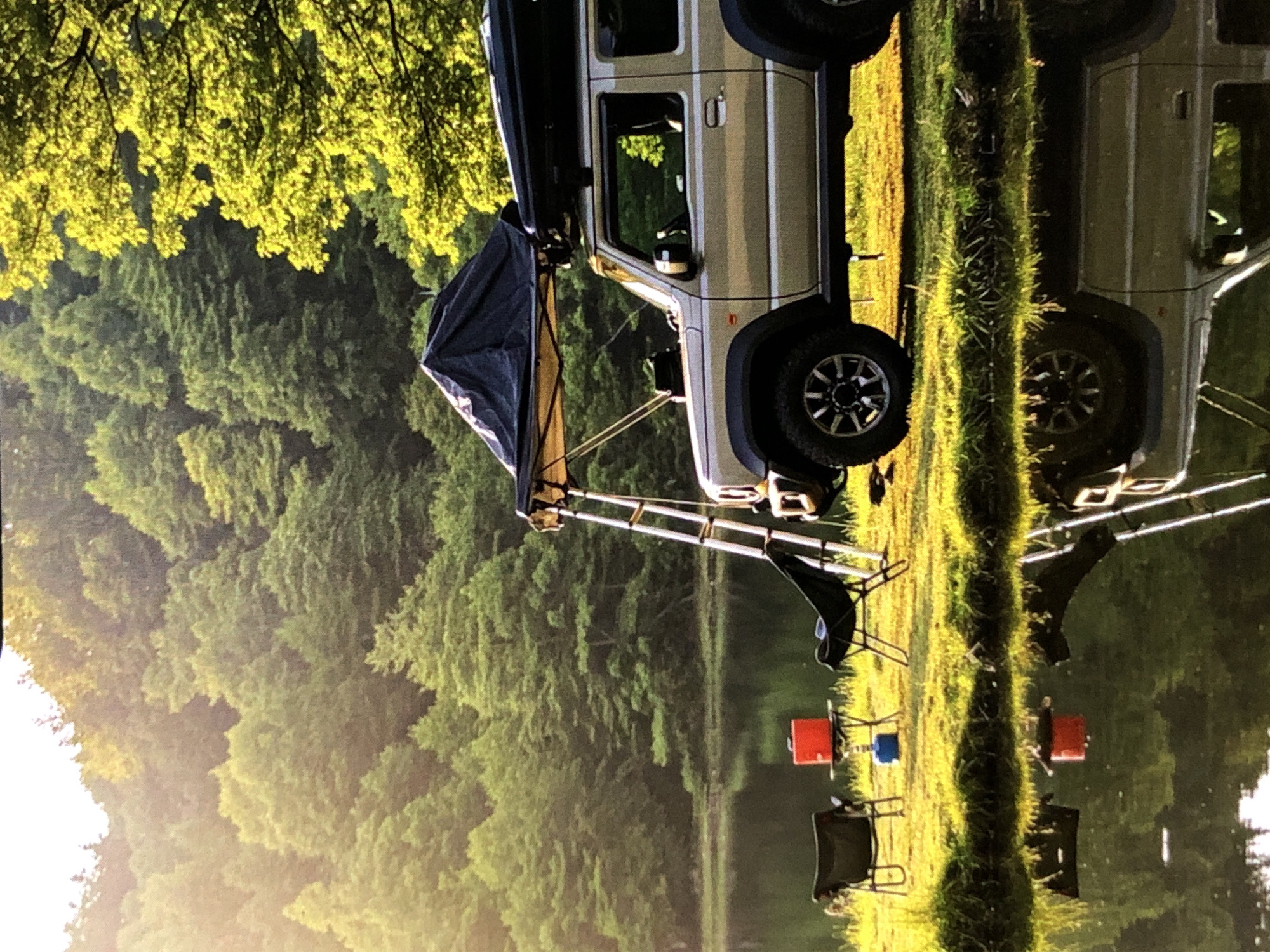 The image shows a silver SUV with a rooftop tent parked on a grassy area next to a calm body of water. Two folding chairs and a small table with a red propane tank are set up near the vehicle. The background consists of a dense forest with lush green trees, bathed in warm, possibly late afternoon or early morning sunlight. The water reflects the trees and the camping setup. The scene depicts an outdoor camping or overlanding scenario. There are no people visible in the image. The location is not identifiable as Bratislava, Slovakia, or any other specific city or region.