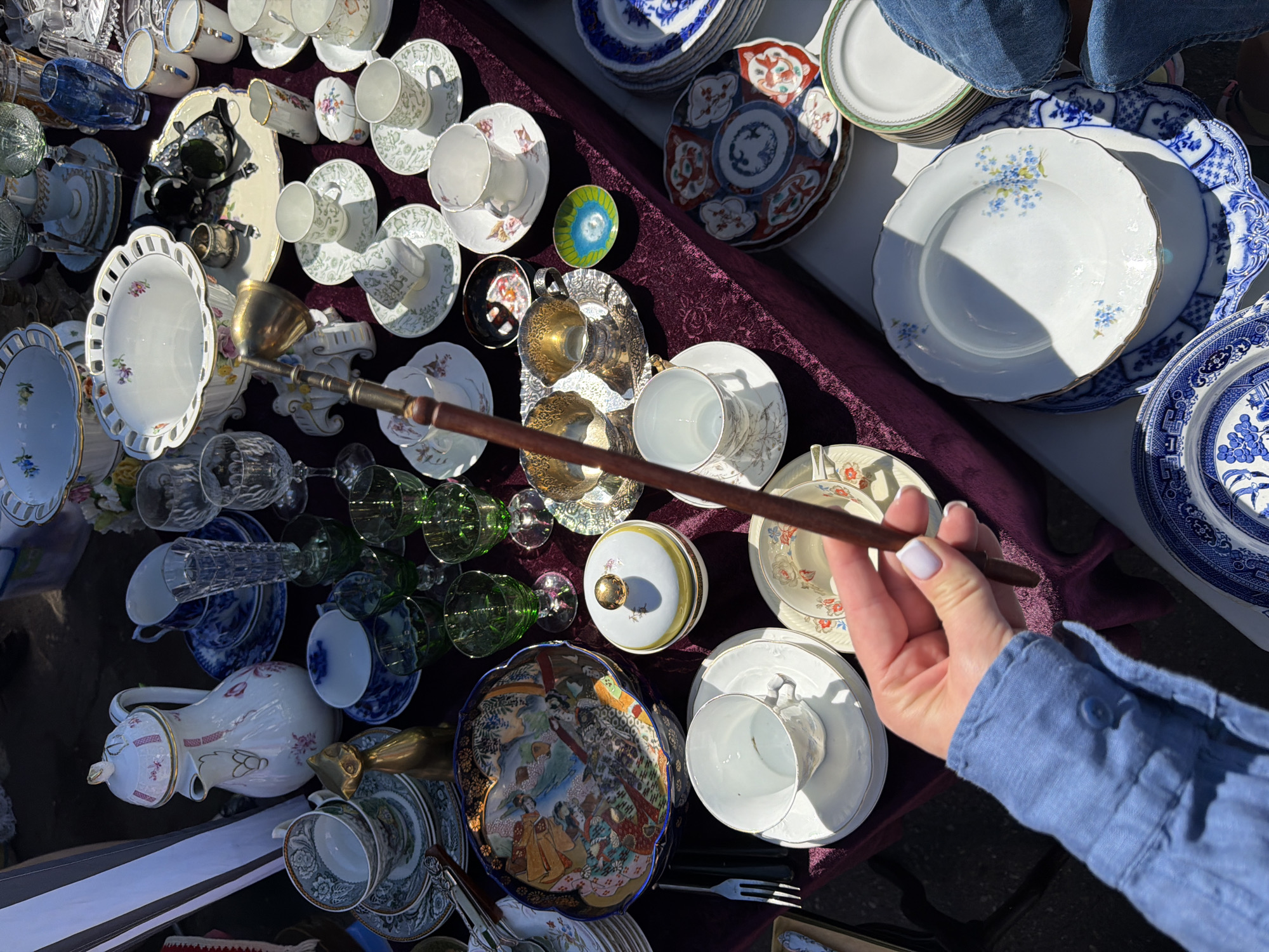The image depicts an outdoor market stall in Riga, Latvia, displaying a dense collection of antique and vintage tableware, primarily porcelain and glass, on a dark purple, possibly velvet, cloth covering a table. Numerous teacups, saucers, and plates in diverse patterns, including floral, blue and white Willowware, and plain white with gold accents, are arranged across the surface. Notable porcelain items include a tall white pitcher with pink floral designs, large decorative bowls with pierced rims and painted motifs, and a deep blue bowl with intricate Asian-style artwork. Glassware consists of various green-stemmed goblets, clear cut-glass stemmed items, and a blue vase. Metal objects include a brass-colored cat figurine, two ornate silver-plated vessels on a small tray, and several dark-handled forks. A person's right hand, with light-colored nail polish, holds a long, slender object with a wooden handle and a brass-colored scoop or bell-shaped end, positioned near the center of the frame. Part of a blue-sleeved arm is visible on the left, and a portion of blue jeans is visible on the lower-right. The scene is illuminated by strong natural sunlight, casting shadows on some items, consistent with an outdoor market environment.
