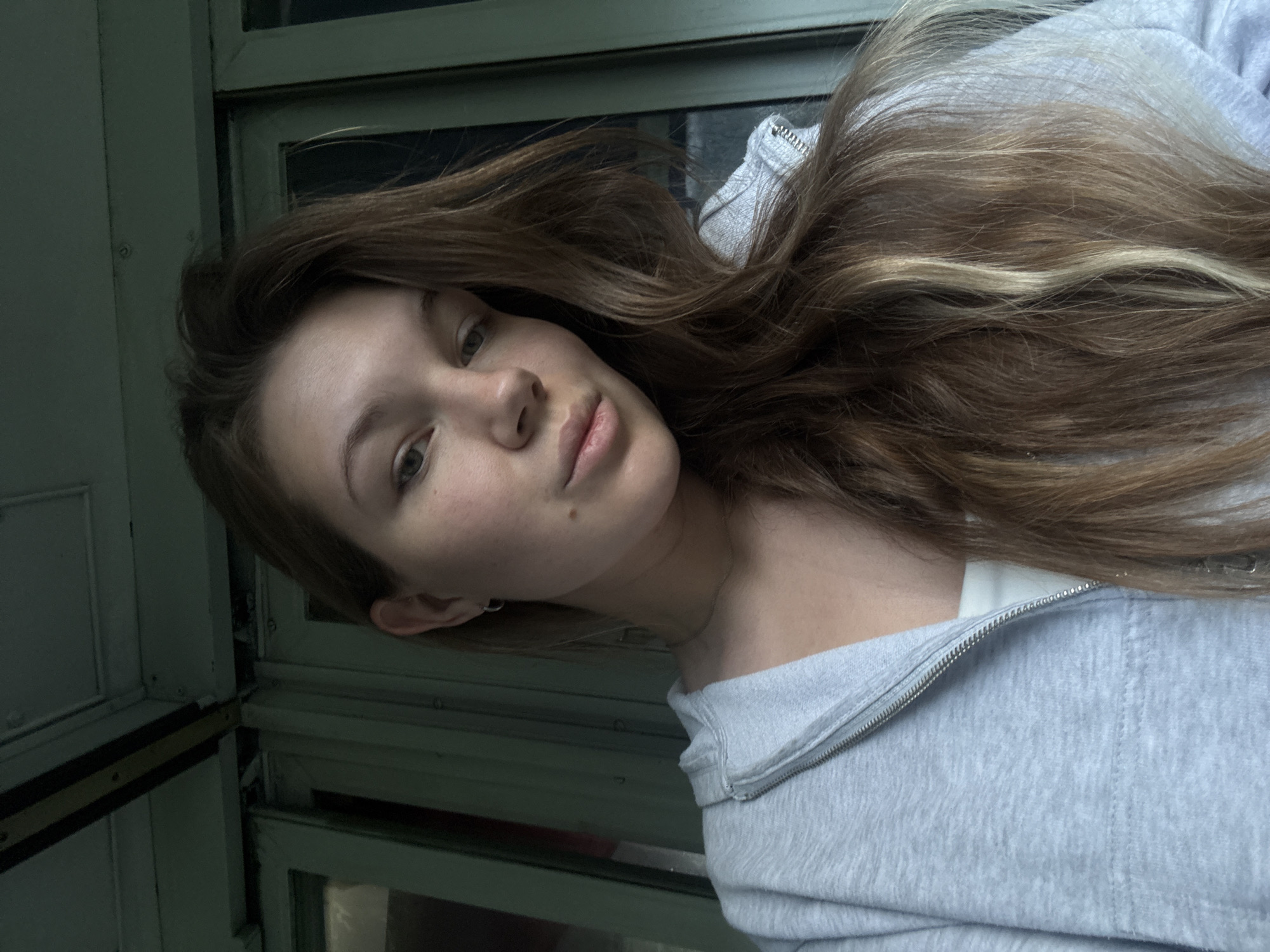 A close-up portrait features a young woman with long, wavy, brown hair streaked with lighter blonde highlights, cascading over her shoulders. She is wearing a light grey hooded sweatshirt with a silver zipper that is partially open, revealing a white undershirt beneath. Her head is gently tilted to her left as she gazes calmly towards the viewer with a neutral expression. Her eyes are light-colored, and a small mole is visible on her chin, below her lower lip. She accessorizes with a thin silver necklace and a small silver hoop earring in her left ear. The background consists of dark green or teal-colored window frames and panels, suggesting the interior of a vehicle like a bus or train car, a common mode of transport in regions such as Bioče, Montenegro. The window structure appears to be segmented with multiple sections or panes. The lighting is soft, indicative of an indoor setting or an overcast day. No discernible text is present in the image.