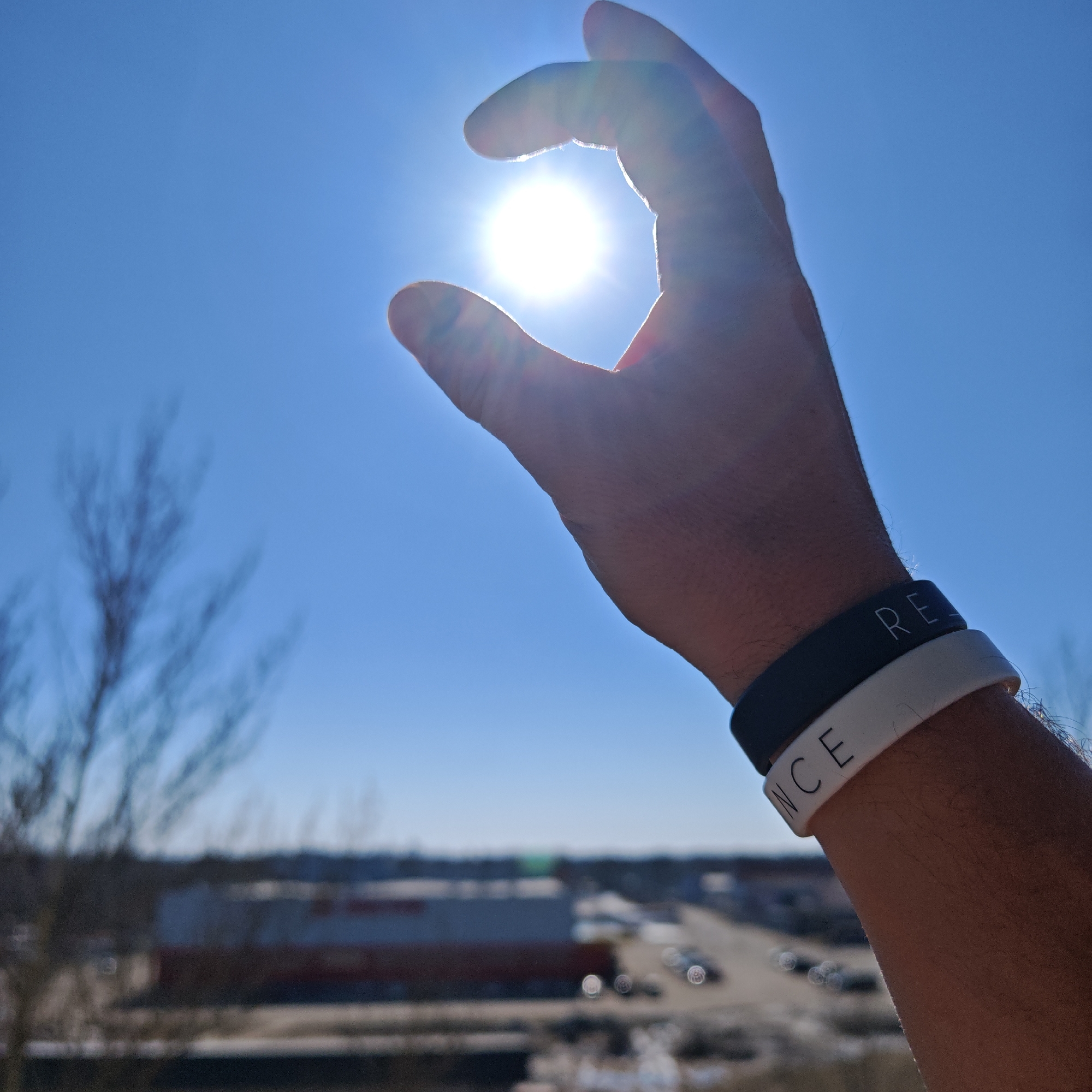 A person's hand is positioned in the foreground, with the sun seemingly held between the thumb and index finger. The wrist wears two silicone bracelets, one black with white lettering reading "RE", and another white with black lettering reading "NCE". The background features a blurred cityscape under a bright blue sky. Bare tree branches are visible to the left. The location is ostensibly Rostov, Russia, but the image itself provides no definitive proof of this other than the text accompanying it.