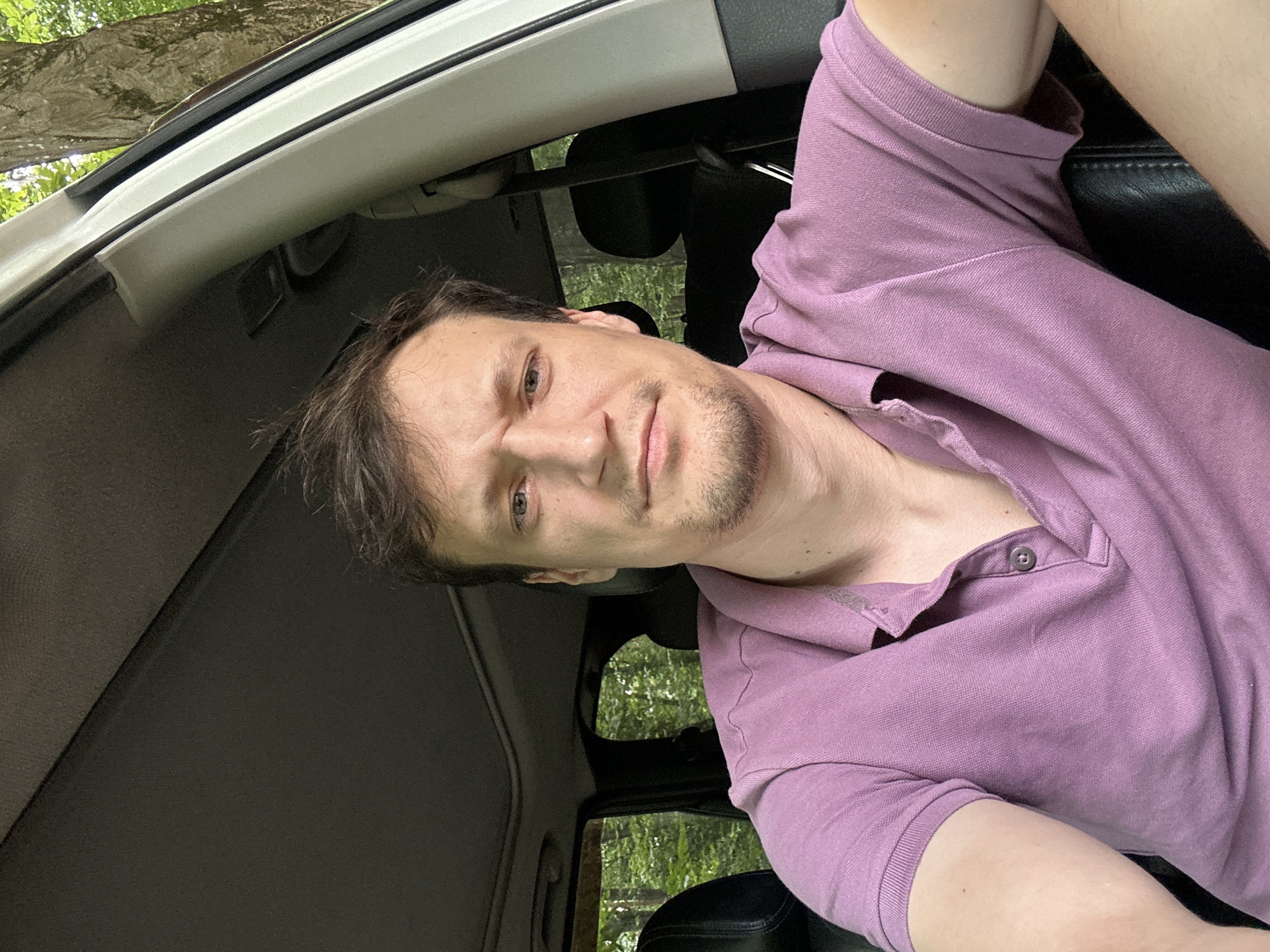 A man with light brown hair and a short beard is sitting in the driver's seat of a white vehicle. He is wearing a purple polo shirt. The interior of the car is visible, including black leather seats and a sunroof. Outside the car, trees and lush green foliage are visible, suggesting a wooded or natural environment. The image appears to be taken during the daytime, with natural light illuminating the scene. The specific location of Shatava, Ukraine, is not visually determinable from the provided image.