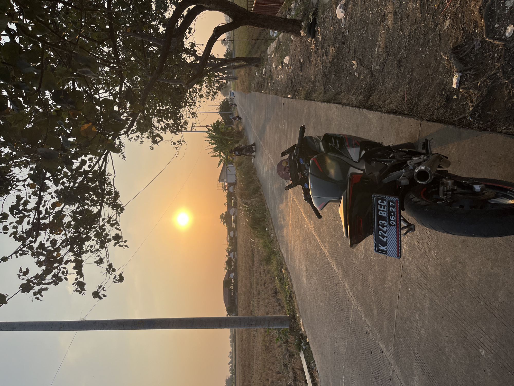 The image captures a scene at dusk on a rural road in Troso, Indonesia. In the foreground, a black sport motorcycle is parked on the concrete road. Its license plate reads "K 4243 BEC" with a date "06-27" below it. The road stretches into the distance, flanked by dry, harvested fields on the left and trees lining the right. A tall utility pole stands to the left of the road. In the background, the setting sun casts a warm glow across the sky, illuminating a few buildings and distant trees. Several other motorcycles and a white car are visible on the road, suggesting moderate traffic. The overall atmosphere is tranquil, with the natural light of sunset dominating the scene.