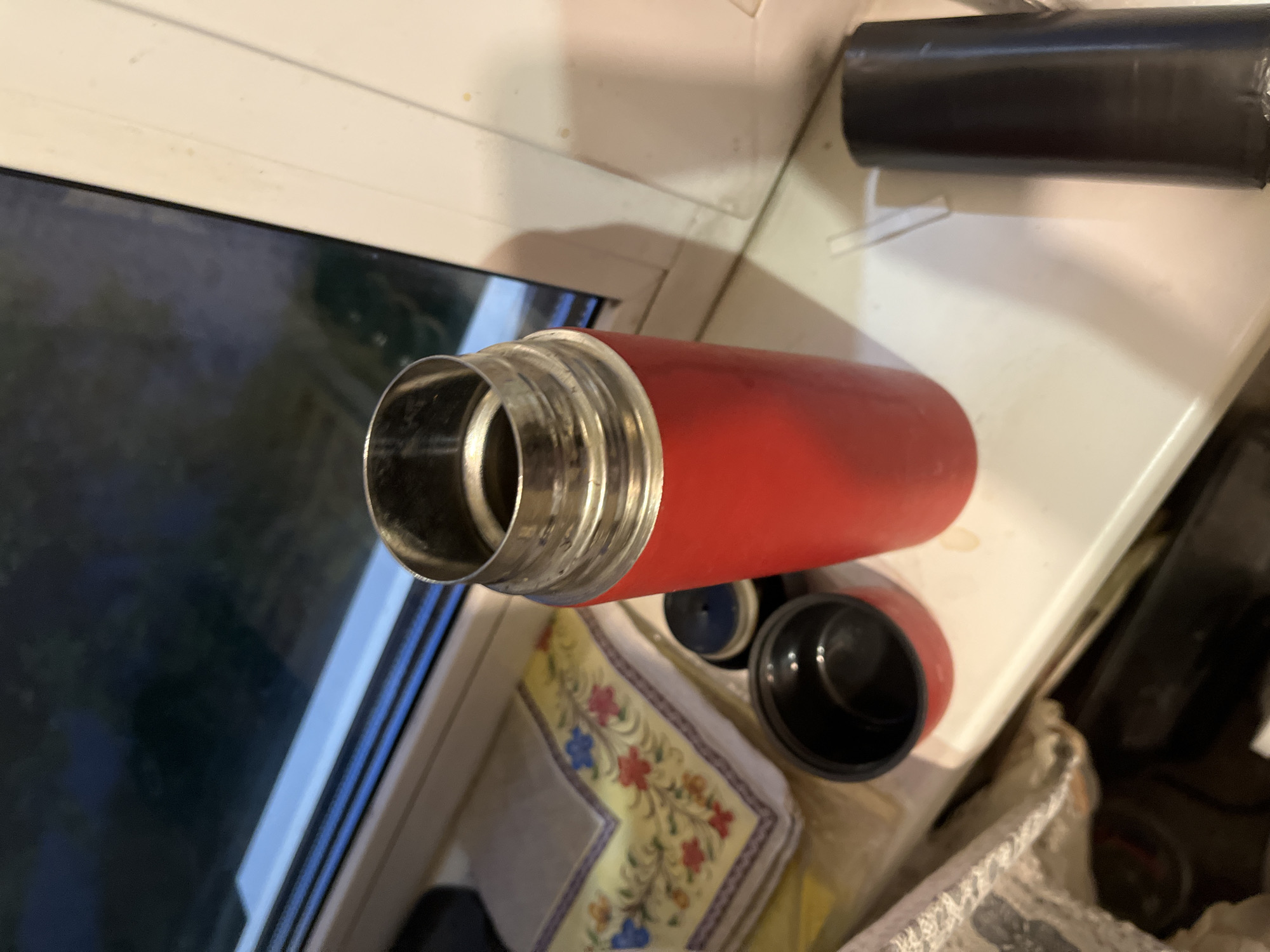 A close-up, high-angle shot shows a red thermos on a white windowsill. The thermos is upright, with its metal lid removed and placed next to it. The lid is black and has a smaller black cap. To the left of the thermos, a folded yellow cloth with a floral pattern is visible. In the background, a window looks out onto a dark, blurry exterior. To the right of the thermos, a dark cylindrical object, possibly a flashlight, is partially visible. The windowsill appears slightly dusty with some faint yellow stains. The scene is indoors, likely in a domestic setting, with the window suggesting it could be in an apartment or house. The context of Minsk, Belarus, is not discernible from the visual elements of the image.
