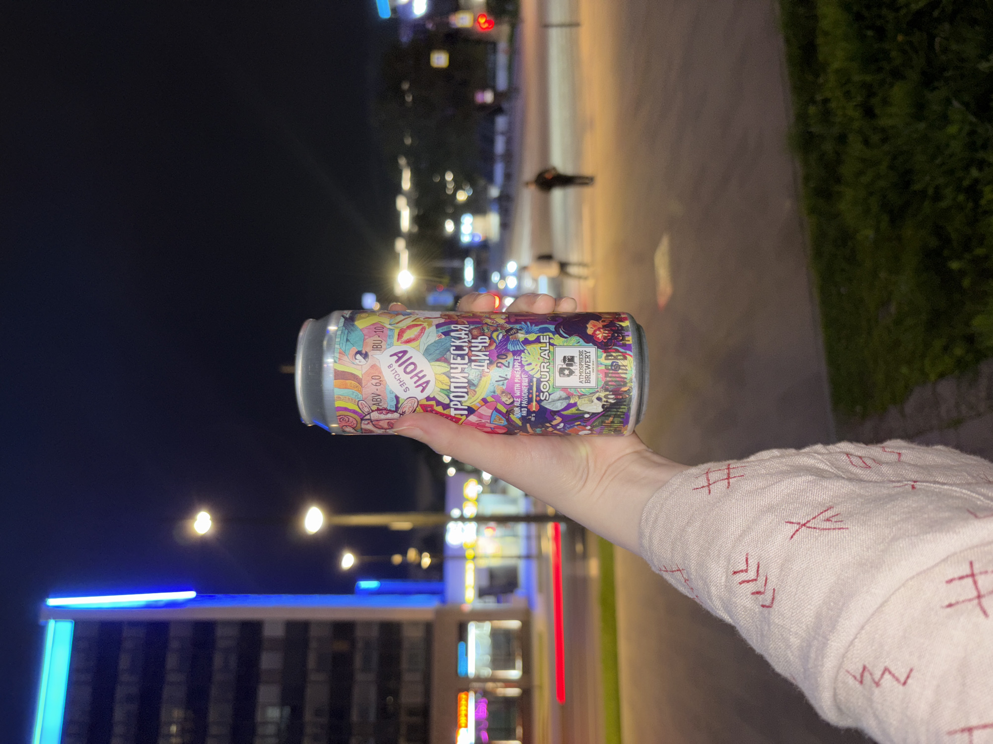 A photograph depicts a person's hand holding a can of "Тропическая Дичь" (Tropical Game) sour ale from Atmosphere Brewery. The label indicates it is V.2.0, brewed with pineapple and passionfruit, with an ABV of 6.0 and 10 IBU. The person is wearing a light-colored sleeve with red embroidered patterns. The background shows an urban scene at night with blurred lights from buildings, streetlights, and moving traffic. Sidewalk and some greenery are also visible. Based on the brewery name on the can, this photograph may have been taken in Perm, Russia.