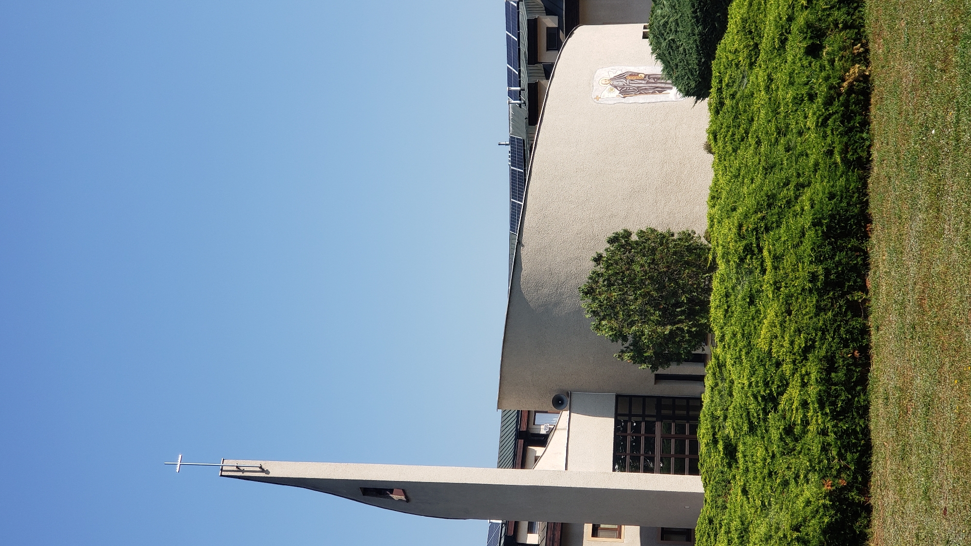 This is a photograph of the Church of the Holy Spirit in Prešov, Slovakia, taken on a clear, sunny day. The church features a modern, curved architectural design with a tall, pointed bell tower topped by a cross. Solar panels are visible on the roof of the adjacent building. A mural depicting a religious figure adorns the curved wall of the church. The foreground is filled with lush green bushes and manicured grass. No people are visible in the image.