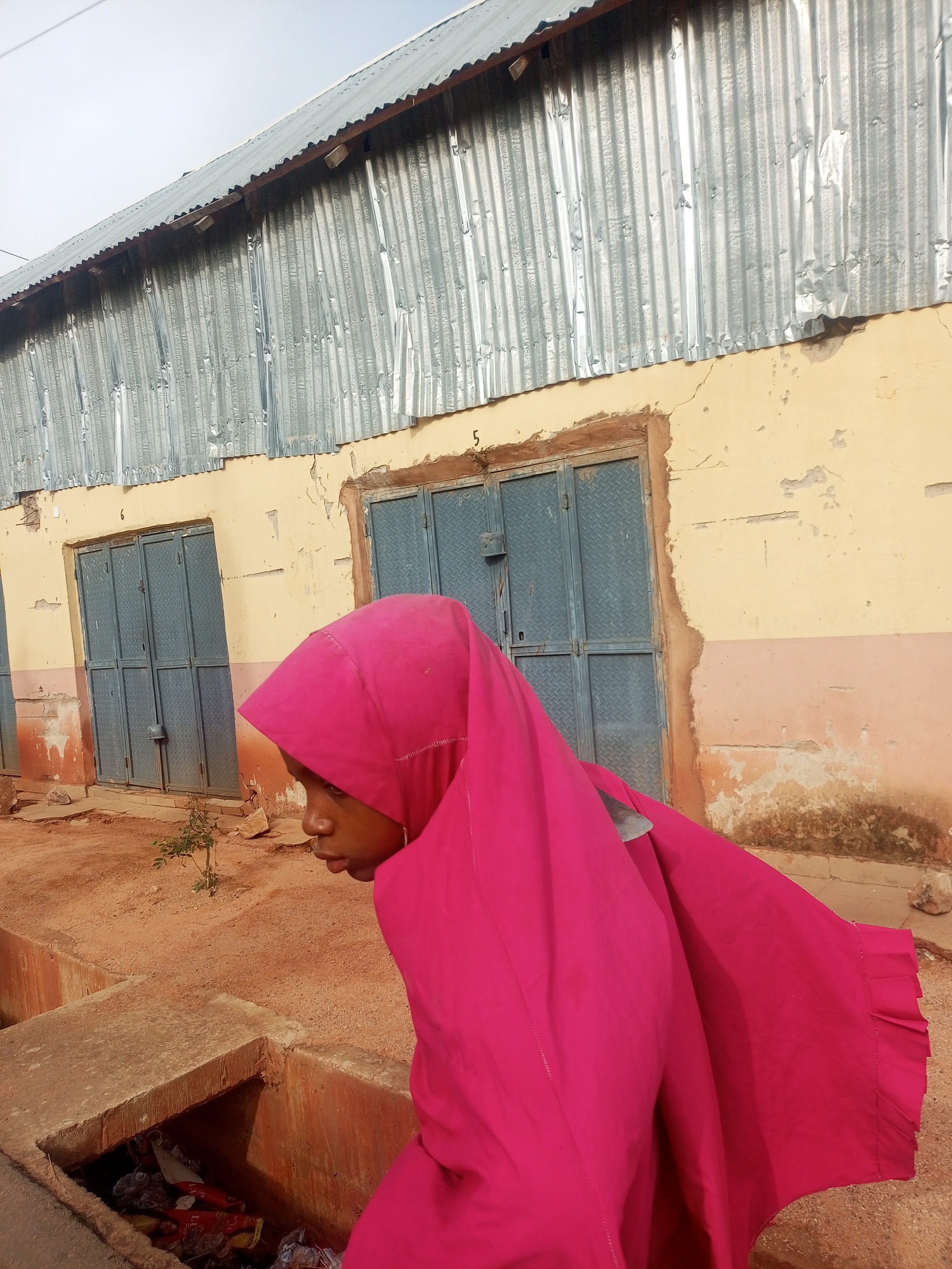 A woman wearing a bright pink headscarf and gown walks past a rundown building with a corrugated metal roof and two metal doors. The building is painted yellow and has some damage. There is debris in the foreground. The location is likely in Tempure, Nigeria.