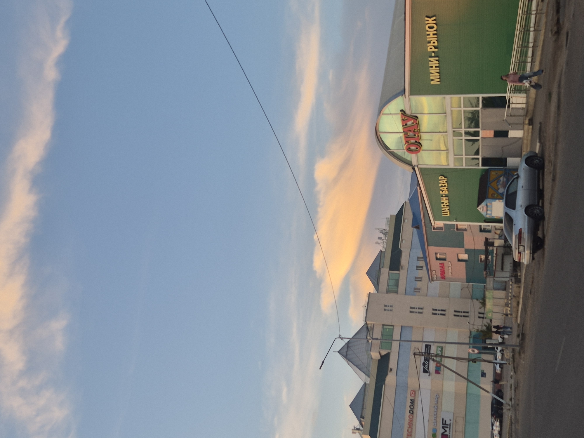 The image depicts a street scene in Kokshetau, Kazakhstan, at what appears to be sunset, given the warm hues in the sky and on the clouds. In the foreground, a silver sedan is parked on the right side of a paved road. Across the road, there are several buildings. The most prominent building on the right is a green structure with an arched entrance and the word "OTAY" in large red letters above it. To the left of the entrance, text in yellow reads "ШАГЫН-БАЗАР" (Shagyn-Bazar), and to the right, "МИНИ-РЫНОК" (Mini-Rynok). A woman in jeans and a pink top is walking on a sidewalk in front of this building, carrying a bag.
Further to the left, a larger, multi-story building is visible, featuring a colorful facade with various signs, including "TECHNODOM.KZ", "MF", and "детский мир" (Detskiy Mir). This building has distinctive pyramid-shaped roofs. A street lamp is positioned in front of this building. Several cars are parked in front of the buildings. The sky is a soft blue with wispy, orange-tinted clouds, suggesting the setting sun. The overall environment appears to be a commercial or mixed-use area of the city.