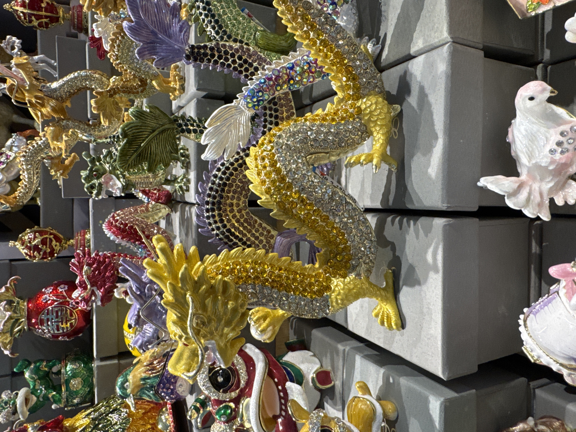 The image shows a display of ornate, bejeweled figurines attached to a wall comprised of grey, stacked boxes. The figurines include dragons in gold, silver, and colorful jewels, as well as other subjects such as birds and decorative eggs. The location is suggested to be in Kuta, Indonesia. The image focuses on the intricate details of the figurines and the repetitive pattern of the grey boxes forming the display wall. There is no visible human presence or specific interactions depicted.
