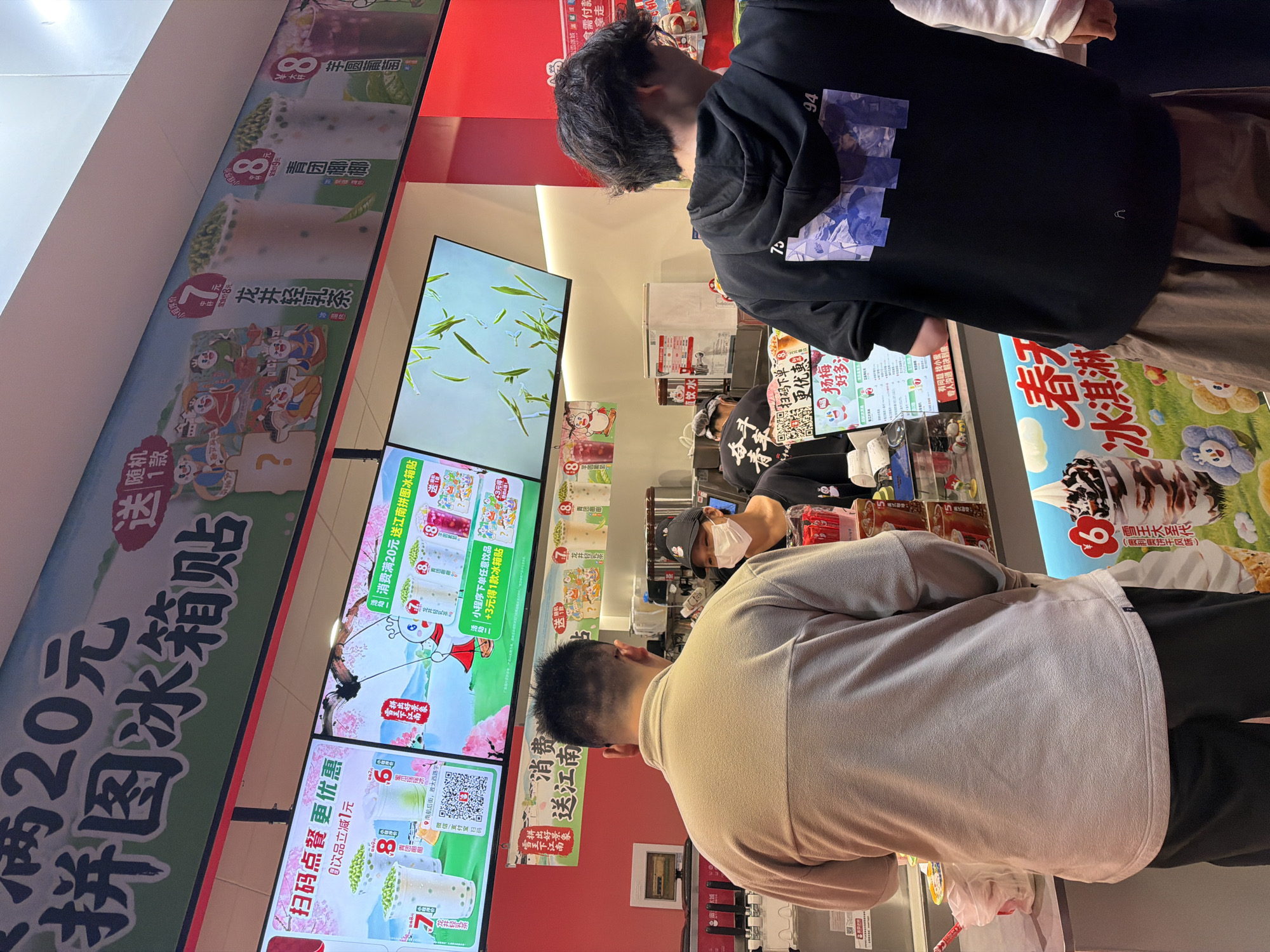 Here is a detailed description of the image:
**Scene**:
The image captures the interior of what appears to be a Chinese bubble tea shop located on NanHanghou Street in the Jiangning District of China. Several people are present, waiting to order or receiving their drinks.
**People**:
The main subjects of the image are the backs of two customers:
* One wears a light brown sweater and black pants.
* The other wears a black hoodie with a graphic design on the back and brown pants.
An employee wearing a face mask and a black hat with a logo is visible behind the counter.
**Objects**:
Menu boards are prominently displayed with images and prices. The menus feature colorful graphics and Chinese text.
* The menu boards are decorated with cartoons and show different types of bubble tea.
* A sign that reads "20 yuan free puzzle refrigerator sticker" is prominently displayed, with promotions written in Chinese.
* There is a display promoting ice cream
* Behind the employee are visible containers for ingredients
**Actions/Interactions**:
The customers are presumably ordering from the employee behind the counter.
**Environment**:
The shop is brightly lit and has a modern design. The walls are painted red and the menu boards are well-lit. The overall impression is that the bubble tea shop is a popular, local venue with a diverse menu.
