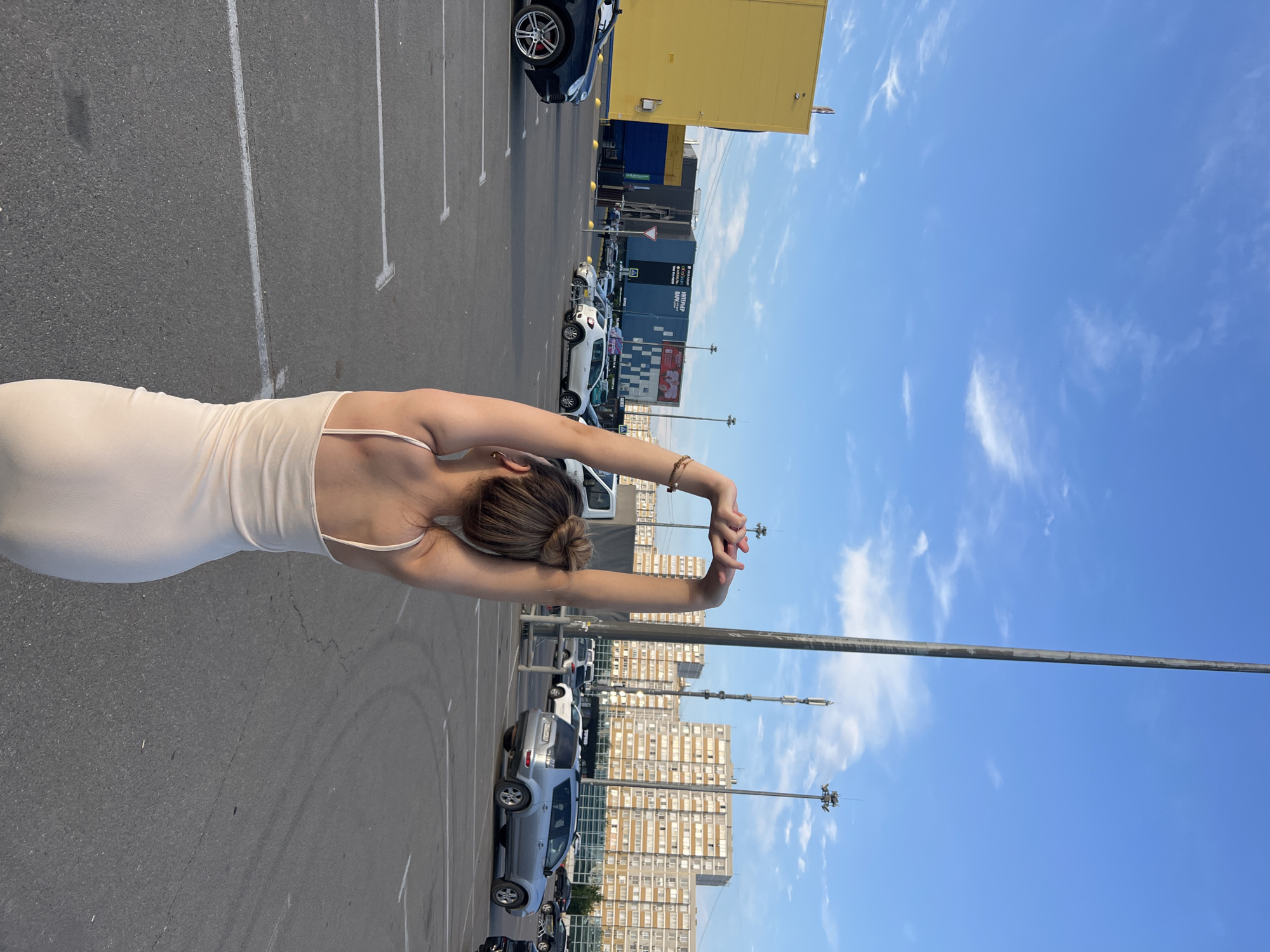 The image depicts a woman with her back to the camera, stretching her arms above her head, in a parking lot. She is wearing a light-colored slip dress. In the background, there are several cars, some buildings, and a tall utility pole. The sky is blue with scattered clouds. The location is a parking lot outside a shopping center in Krasnodar, Russia, recognizable by the signage on the buildings which appears to be in Cyrillic script and includes text like "INTERIOR PARK". The overall scene suggests a commercial area during daylight hours, likely late afternoon or early evening due to the lighting.