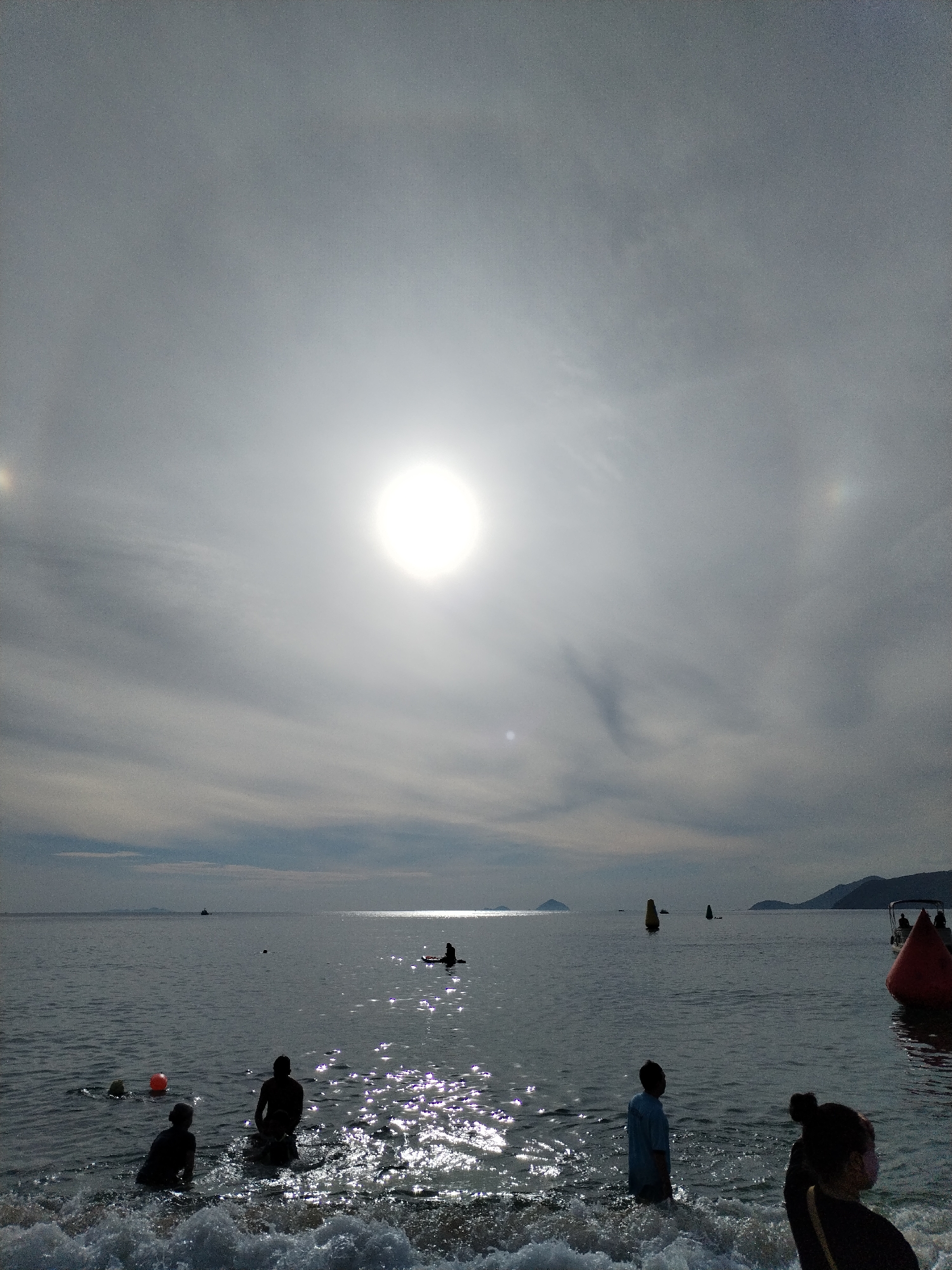 This is a photograph taken at a beach in Nha Trang, Vietnam, under a bright, slightly hazy sun that is high in the sky. The sun appears as a strong, luminous disc. The sky is filled with a mix of clouds, with some areas appearing brighter due to the sun's glare.
The scene depicts the ocean, with gentle waves breaking at the shore. The water is dark and reflects the sunlight, creating a shimmering effect.
In the foreground, several people are visible in the shallow water and at the water's edge. To the left, there are two figures partially submerged in the water, with a small orange buoy nearby. Further into the water, another figure is seated. To the right, two more figures stand in the water, one appearing to be a child in a light blue shirt, facing away from the camera. A person with their back to the camera is also visible on the right, with their hair styled in a bun.
In the middle ground of the ocean, a person is seen sitting on a paddleboard. Various buoys are scattered across the water, including a large red conical buoy on the far right, and yellow buoys further out. A small boat with people on board is visible in the distance on the right. In the far distance, hills and mountains form the coastline of Nha Trang. The overall impression is a serene beach scene on a sunny day.