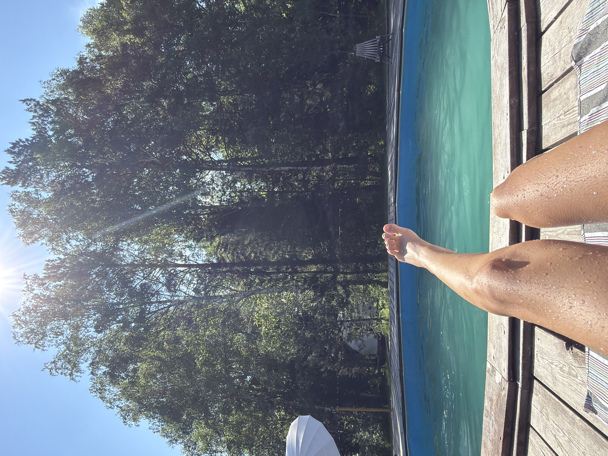 This is a first-person perspective, eye-level, outdoor, close-up shot of a person's legs and feet resting on a wooden deck next to a bright blue swimming pool. The water in the pool is calm with slight ripples. The person's skin appears tanned and has water droplets on it. In the background, a dense forest of green trees stretches across the horizon under a clear blue sky with a bright sun causing lens flare. A white umbrella is visible to the left, and a wooden lounge chair is situated to the right, both near the pool. The location is not discernible from the image and cannot be identified as Минино, Russia.
