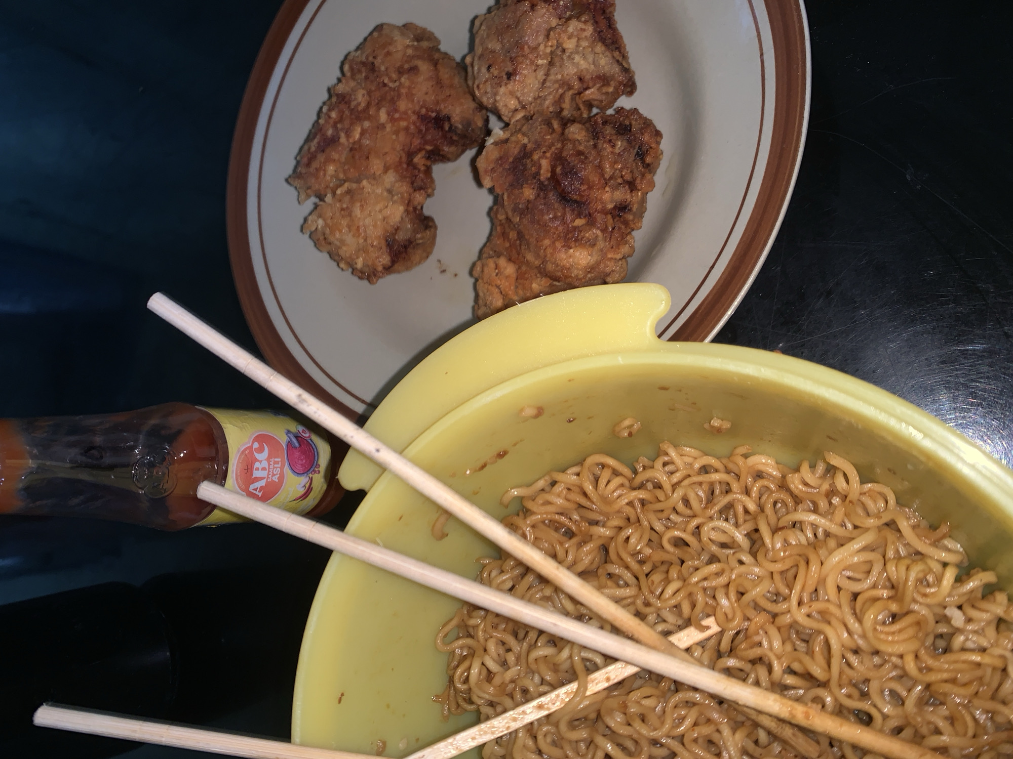 A close-up, overhead shot shows a yellow bowl filled with instant noodles, a plate of fried chicken, and a bottle of ABC Sambal Asli hot sauce. The noodles are cooked and appear to be mixed with a sauce, with three wooden chopsticks resting on top. The fried chicken, three pieces visible, are golden brown and crispy. The ABC Sambal Asli bottle, with its distinct red and yellow label, is positioned behind the bowl of noodles. The scene is set on a dark, reflective surface, possibly a table. The lighting is dim, casting shadows and highlighting the textures of the food. This image depicts a casual meal, likely prepared or consumed in a residential setting within Sungai Meriam, Indonesia.