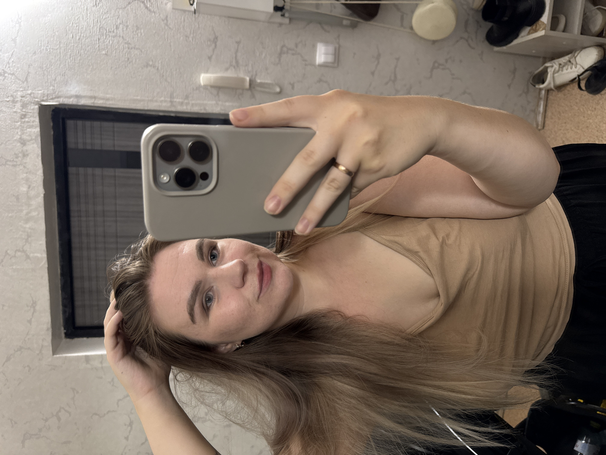 A woman is taking a selfie indoors. She is holding a smartphone with a gray case, and her hand has a gold ring. She is wearing a tan top. The background includes a patterned wall, a window with horizontal blinds, and a shelf with various items like shoes and a white sphere. The location may be in Chaikovsky, Russia.
