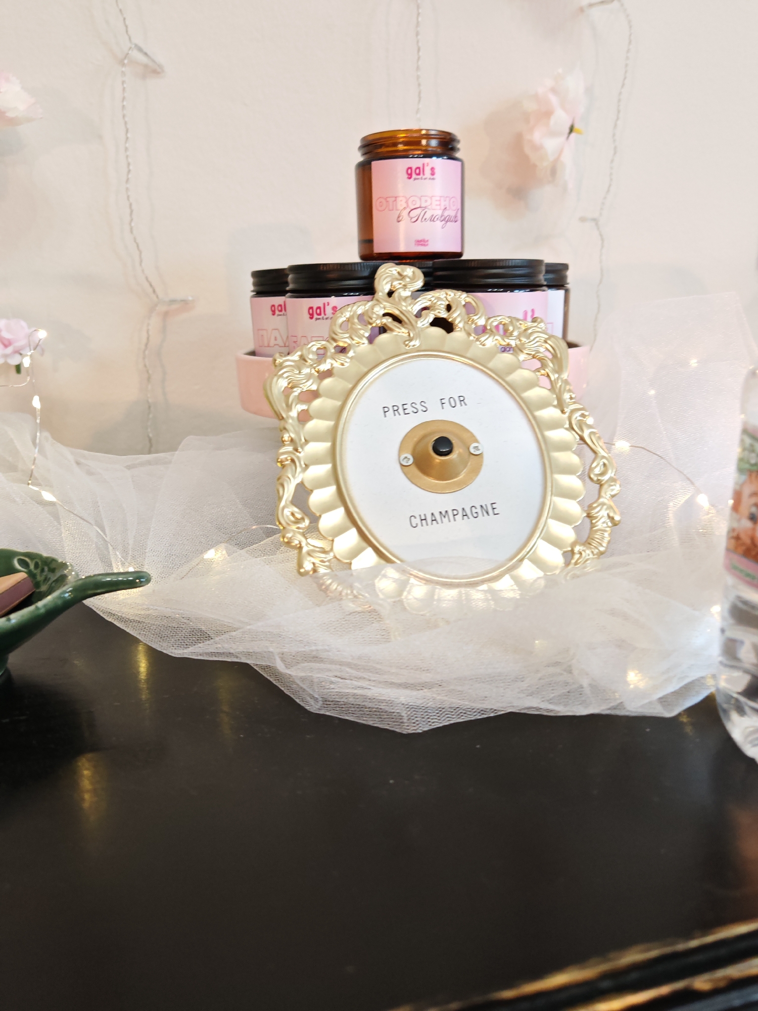 A close-up, eye-level shot shows a decorative arrangement on a dark, reflective surface, likely a table. In the foreground, a small, green, leaf-shaped dish holds several brown objects, possibly chocolates or candies. Behind the dish, a white, sheer fabric is draped, adorned with small, twinkling LED lights. Centered in the frame is an ornate, gold-colored oval frame containing a white plaque. The plaque has black text reading "PRESS FOR CHAMPAGNE" and a gold button with a black center, suggesting a novelty doorbell or call button. Stacked behind the frame are three pink jars with black lids, labeled with "gal's" in pink lettering. The topmost jar is slightly tilted and has additional pink text on its label, which appears to be in Cyrillic. The background is a pale, textured wall, with faint, decorative pink flowers and string lights visible. The overall aesthetic is whimsical and celebratory. The mention of Plovdiv, Bulgaria, is not supported by visual evidence in the image itself, as there are no identifiable landmarks or signs indicating a specific city or location within Plovdiv.