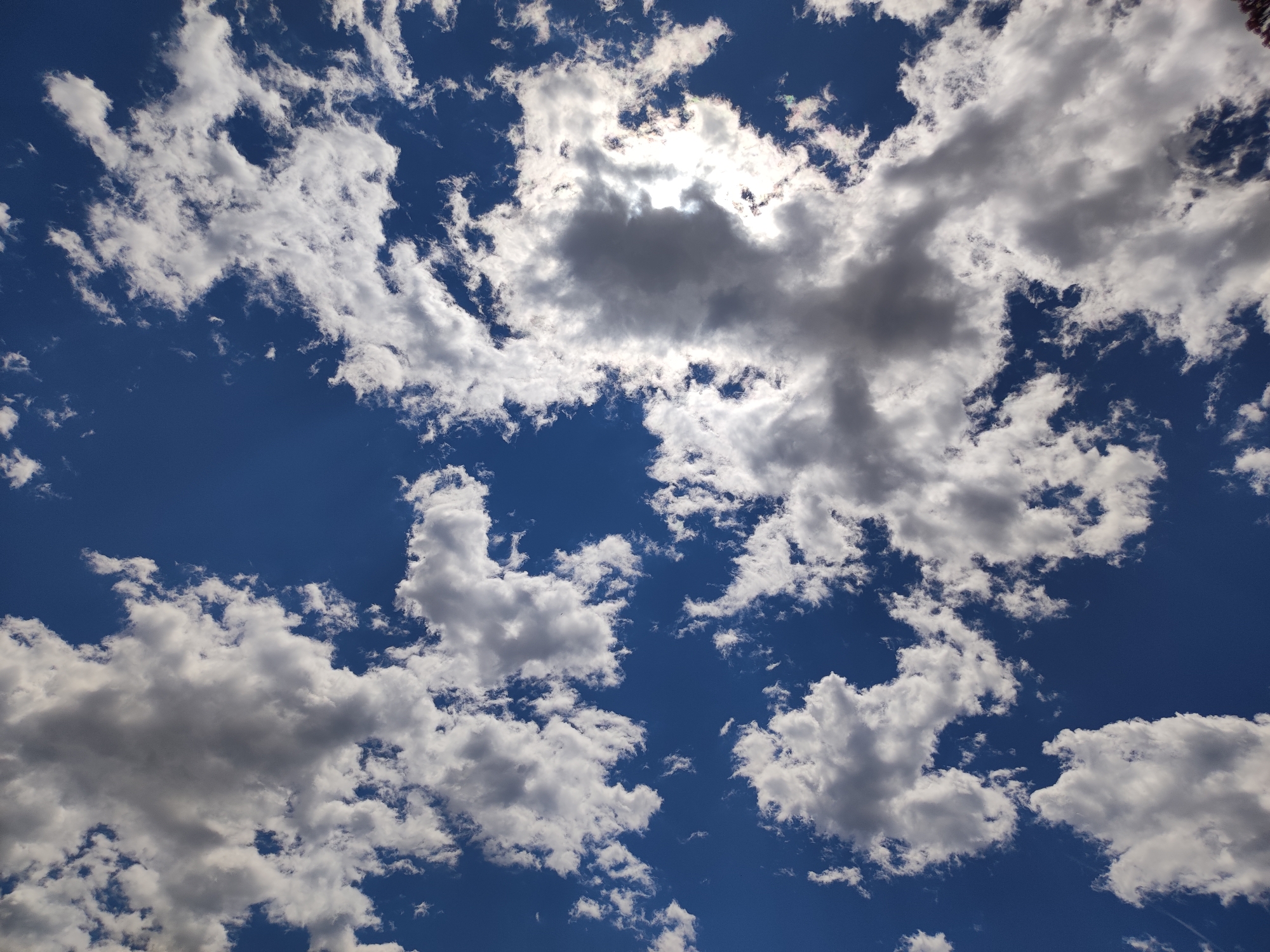 The image displays a bright blue sky filled with a variety of clouds, ranging from fluffy white cumulus to darker, more shadowed formations. The sun is partially obscured by a large, dense cloud in the upper center of the frame, creating a bright glare and casting shadows on surrounding clouds. The clouds are dispersed across the sky, with some areas showing patches of clear blue sky. The scene is a natural outdoor environment, indicative of a daytime sky above Meran - Merano, Italy. There are no people, subjects, or objects visible in the image other than the clouds and sky.