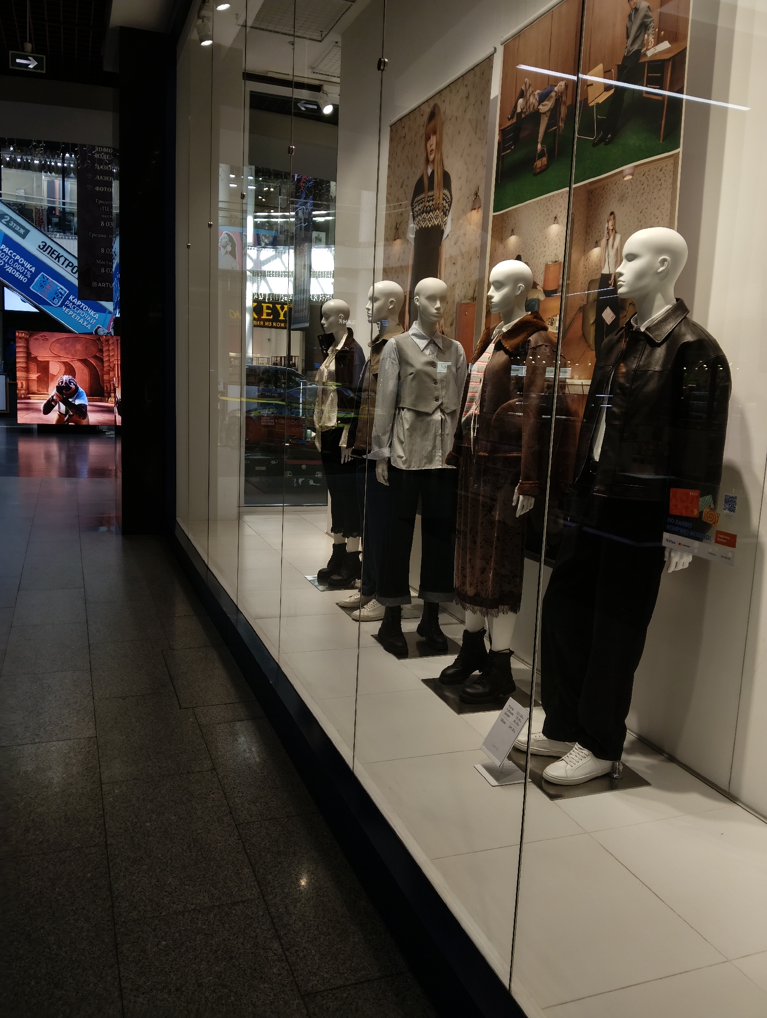 The image shows a storefront window display featuring several mannequins dressed in fashionable clothing. The mannequins are arranged in a row, each wearing a different outfit, including jackets, shirts, and pants. The display includes large posters in the background with images of models wearing similar clothing.
The storefront is located in the city of Hrodna, Belarus, as indicated by a sign that reads "Гродно" (Hrodna) and other Russian text which appears to be advertising or store information. The floor of the storefront is tiled with a light color, while the flooring outside the display is a darker, speckled tile. Reflections of the interior of the store and the exterior environment are visible on the glass window.
On the left side of the image, a large digital screen displays what appears to be an advertisement for "ЭЛЕКТРО" (Electro) and other Russian text. The screen shows an animated scene with cartoon characters, likely from a movie or show. The surrounding area appears to be a shopping mall or a similar indoor commercial space, with a corridor visible on the left.
