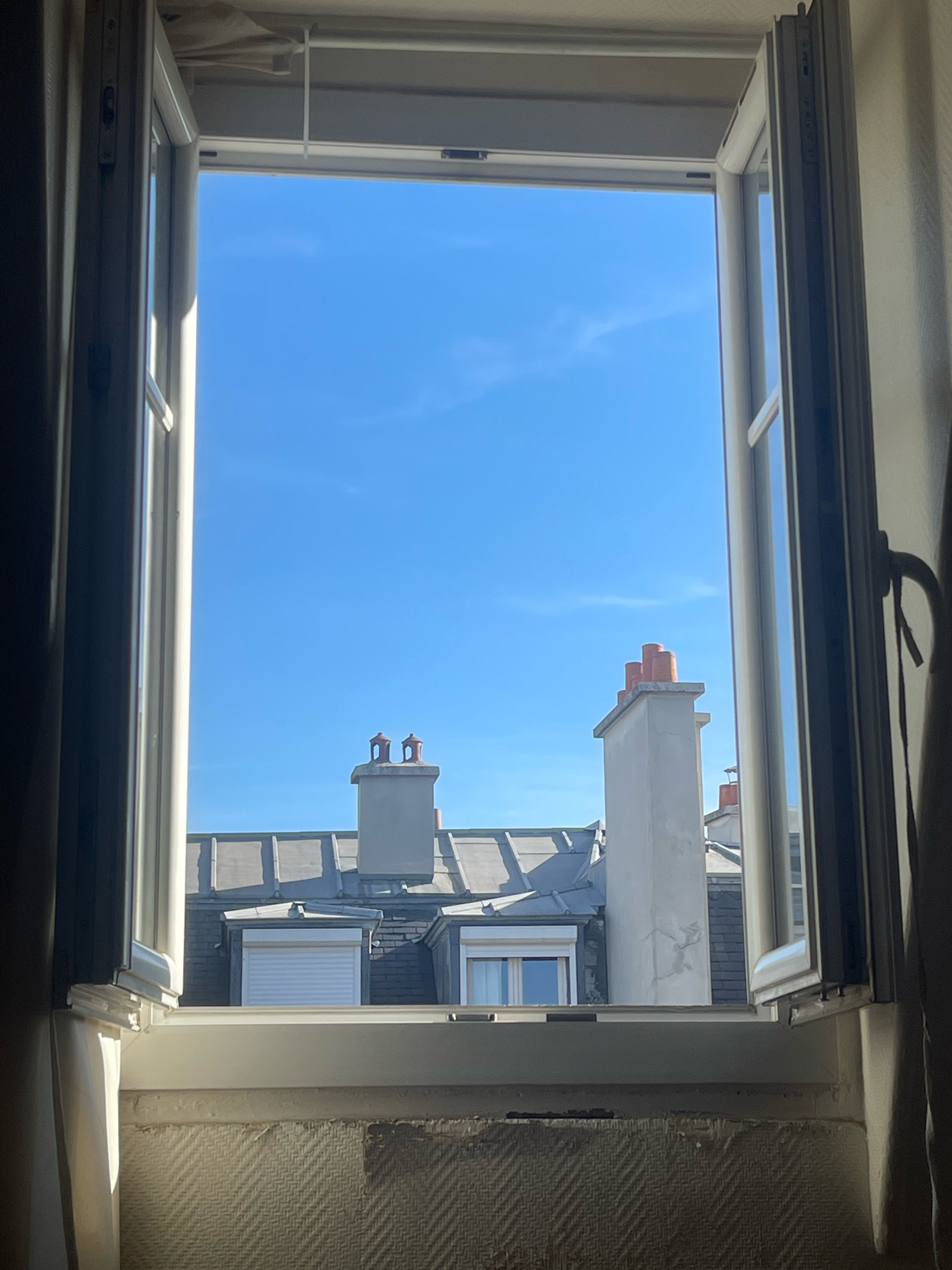 The image captures a view through an open window in Paris, France, looking out onto a residential rooftop scene under a clear blue sky. The window frame is white and appears to be modern. One pane is open inward.
The view beyond the window reveals the distinctive architecture of Parisian rooftops. There are several chimneys visible. One prominent chimney, on the right side of the frame, is tall and white with a light-colored cap and appears to have some fine cracks on its surface. To its left, another, shorter chimney is grey with a similar cap, and has two smaller chimney pots on top. Further to the left, another chimney, mostly obscured, is visible with red chimney pots.
The roofs are clad in grey slate tiles, with a series of mansard roofs and dormer windows. One dormer window, in the center, has a white roller shutter drawn down. Another dormer window to its right shows a clear glass pane, reflecting the sky. The overall impression is of a densely built urban environment.
In the foreground, the bottom of the window frame is visible, with a textured, wallpapered wall below. The wallpaper has a repeating chevron pattern in a neutral color. Sunlight streams in from the left, casting shadows on the wall and the window frame. The scene suggests a bright, sunny day in the city. No people are visible in the image.