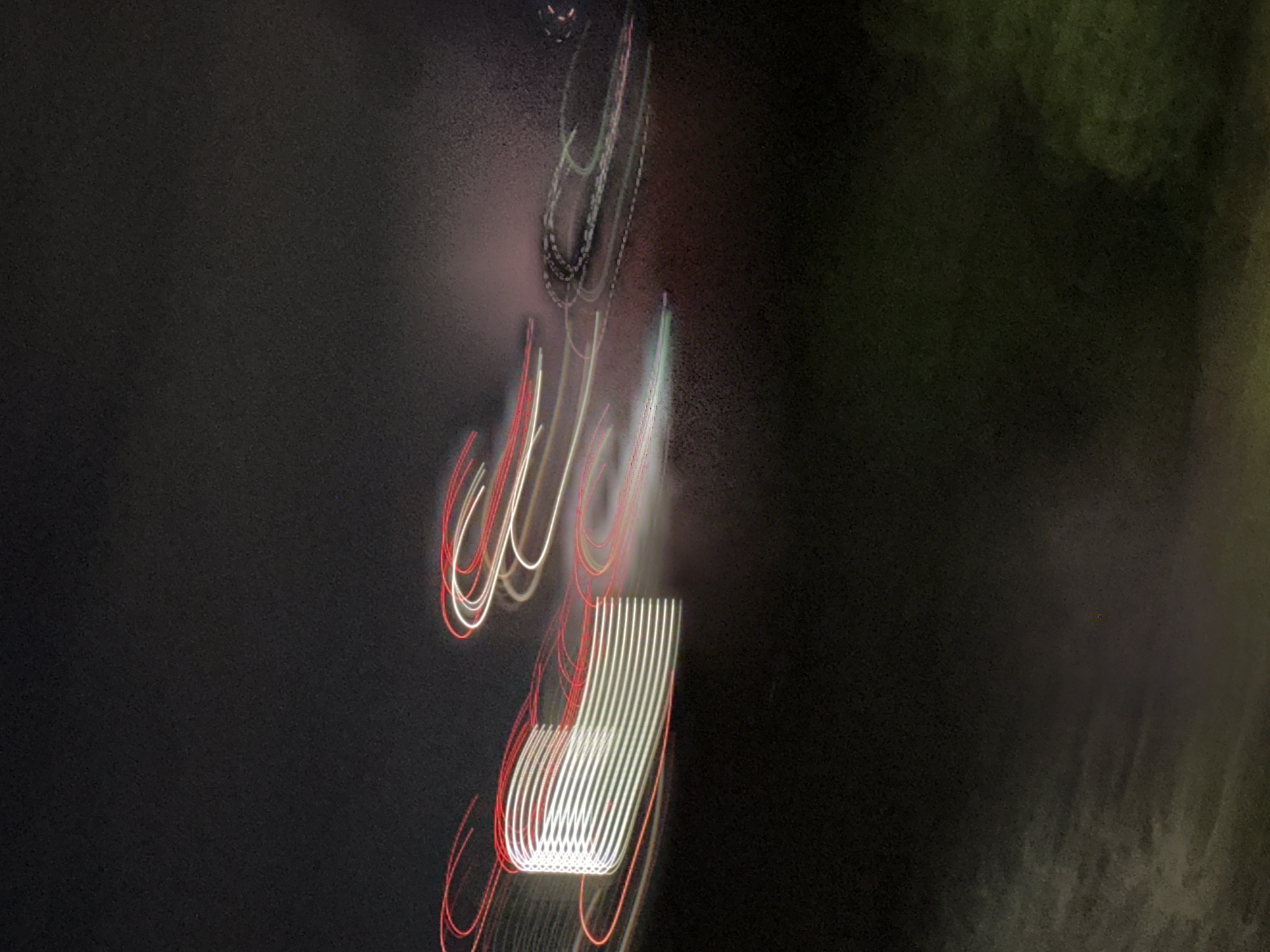 The image shows a blurred, nighttime photograph in Nizhny Novgorod, Russia, featuring light trails from moving objects. The most prominent light trails are a series of horizontal, parallel white lines with red edges, suggesting a moving vehicle or object with illuminated edges. Above this, there are blurry trails of red and white curved lines. Higher in the frame, there are faint, circular light trails in white and green. The background is dark, with the left side appearing darker than the right, which has a slight greenish tint, potentially due to vegetation or ambient light. The photograph appears to be taken with a slow shutter speed, resulting in the blurred light trails.