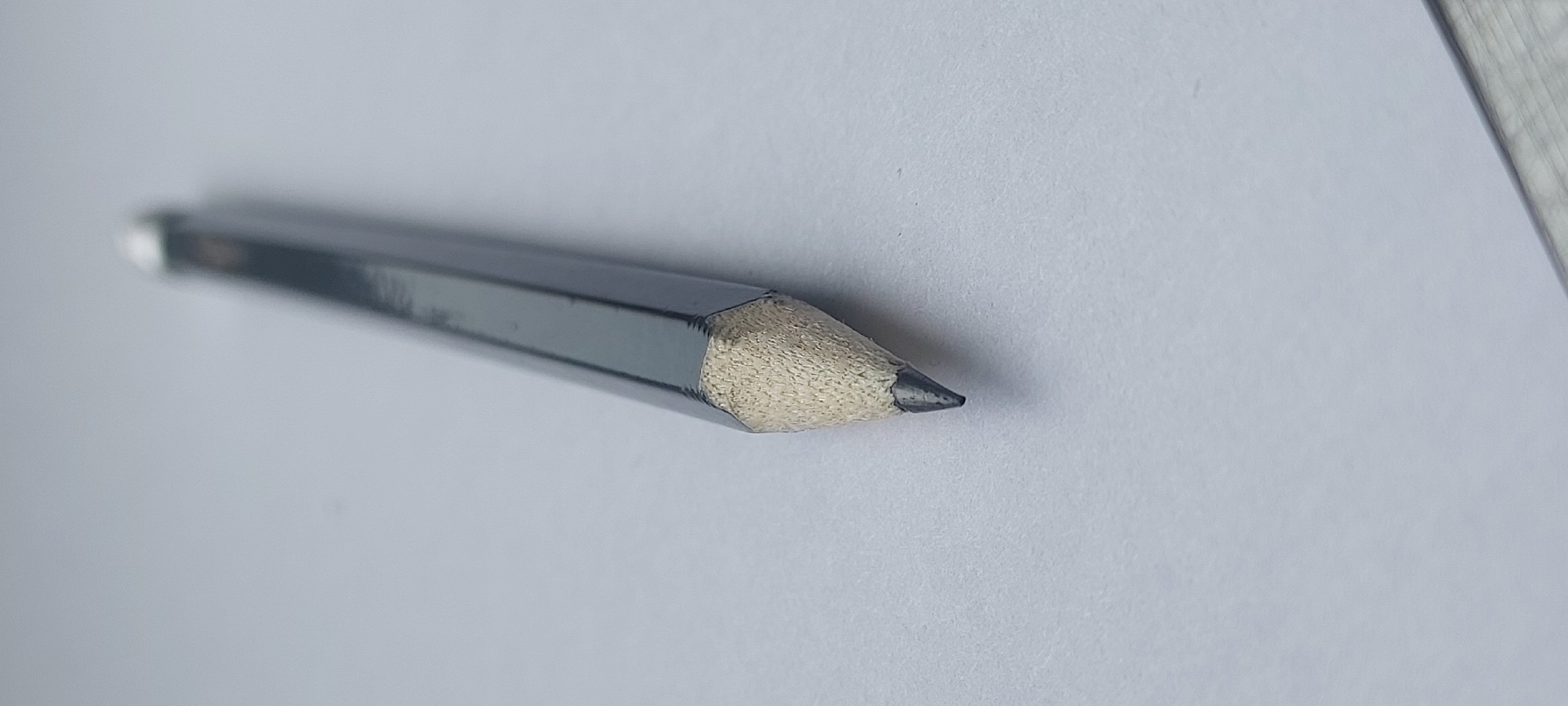 A sharpened graphite pencil with a hexagonal gray barrel rests diagonally on a white, textured surface, likely paper. The pencil's tip is pointed and dark, with visible graphite. The wood casing around the graphite is exposed and appears light brown. The background is a plain white, with a slight shadow cast by the pencil. There are no people or other objects visible in the frame. The image is a close-up, with shallow depth of field, blurring the background. The scene does not depict any recognizable urban or natural landscape elements, and therefore, its location within Storozhevaya-2, Russia, or any other city, cannot be determined from the visual information provided.