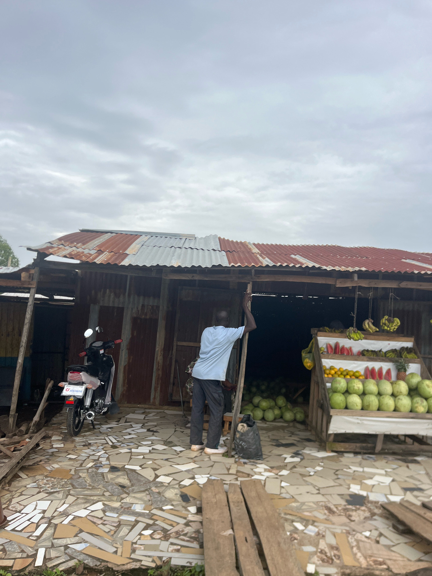 This image depicts an outdoor market stall in Barakin Akawo, Nigeria, under an overcast sky. A man in a light blue t-shirt and dark pants stands near the entrance of the stall, leaning against a wooden post. The stall itself is constructed with corrugated metal and wood, featuring a weathered, rusted roof. In front of the stall, a variety of fruits are displayed on shelves: watermelons, oranges, and bananas are visible, along with slices of watermelon and a pineapple. Behind the stall, a large quantity of green fruits, likely watermelons, are stacked. To the left of the stall, a black motorcycle is parked. The ground in front of the stall is paved with broken tiles of various colors and patterns. A black trash bag is visible near the man. The overall scene suggests a typical roadside market environment.