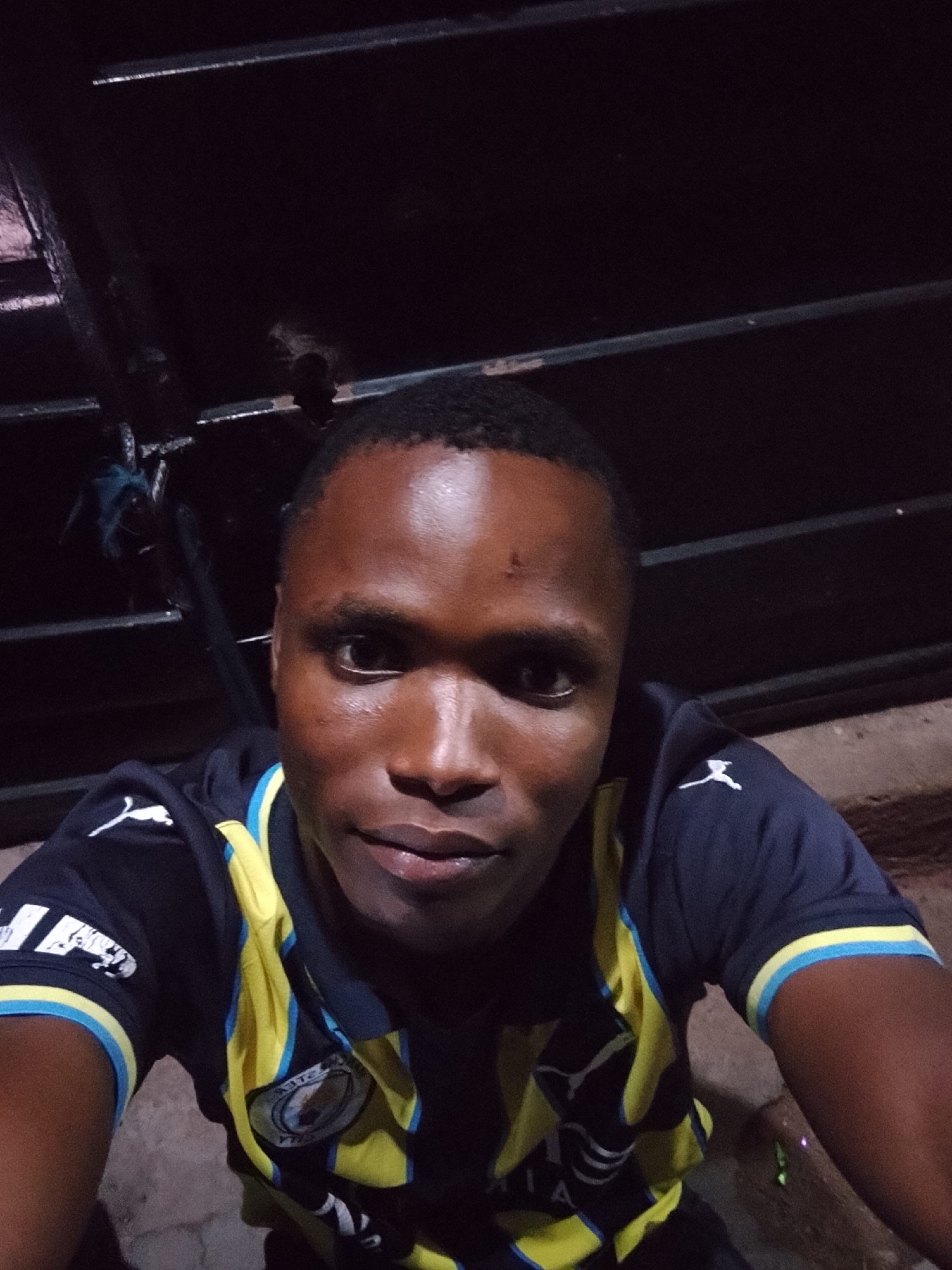 A dark-skinned man with short hair is taking a selfie in a dimly lit, outdoor environment, possibly at night. He is wearing a navy blue t-shirt with yellow and light blue accents. The t-shirt has a Puma logo on the left shoulder. In the background, there are dark, horizontal structures that appear to be metal bars or grating, possibly part of a fence or a railing. The lighting is low, creating shadows and highlighting the man's face. The ground appears to be rough, possibly concrete or dirt. The man is looking directly at the camera.