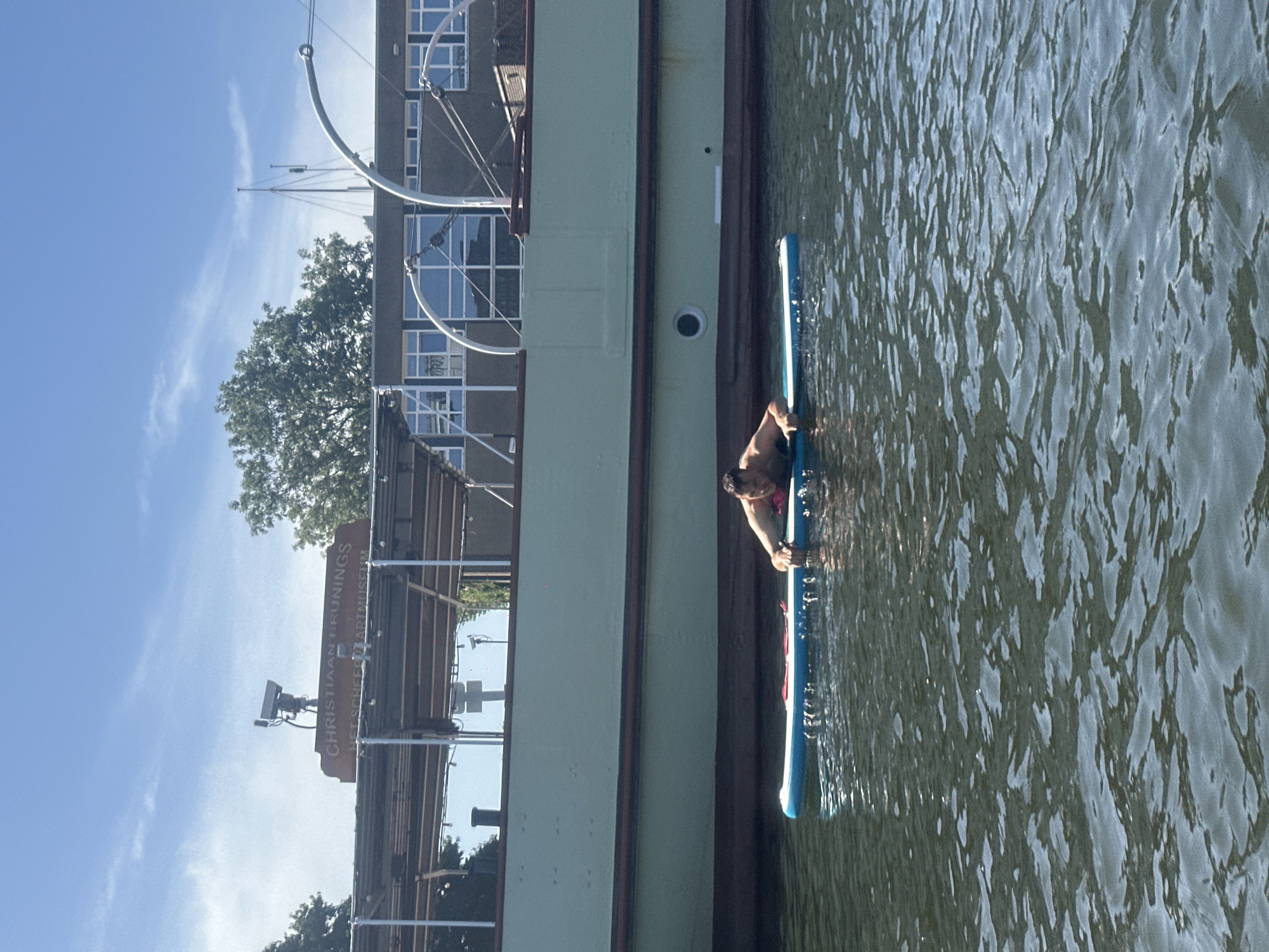 A man is paddleboarding on the water in front of a large, light green ship. The ship has a dark brown trim and a dark brown hull. Above the ship, there is a building with many windows and a sign that reads "CHRISTIAAN BRUNINGS HET SCHEEPVAARTMUSEUM". The sky is blue with some white clouds. The water is dark and rippled, reflecting the sunlight. The man is wearing dark shorts and appears to be holding onto the paddleboard with his hands. The paddleboard is blue and white.