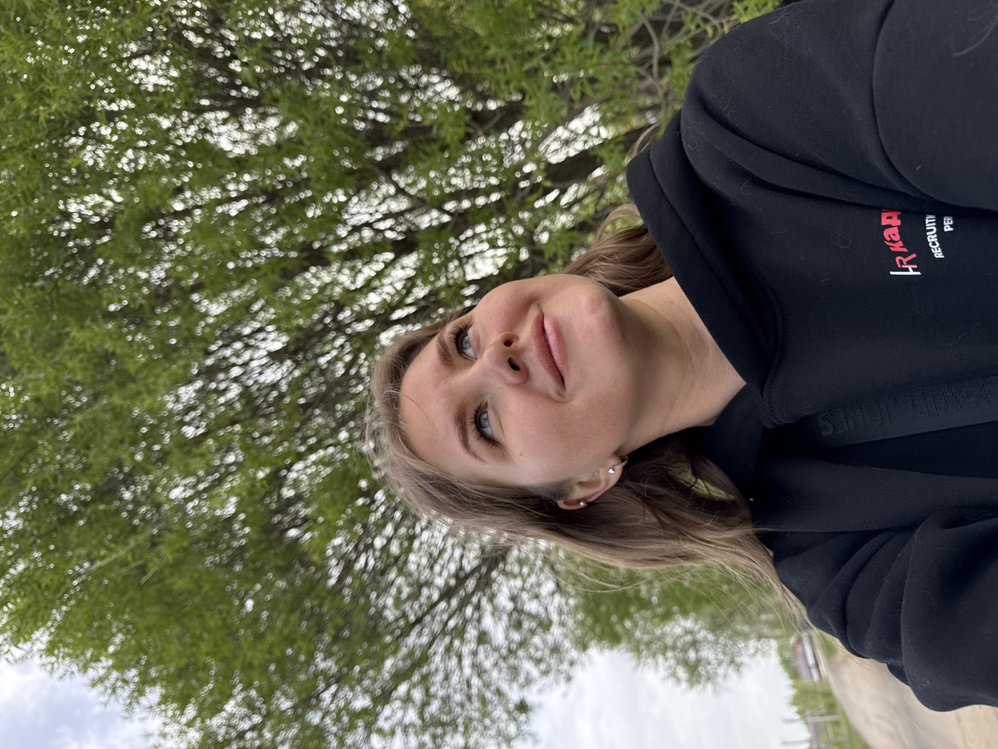 A young woman is standing outside in Горный, Russia. She is wearing a black hoodie with red and white lettering on the sleeve. A tree with green leaves is behind her. The weather appears overcast.