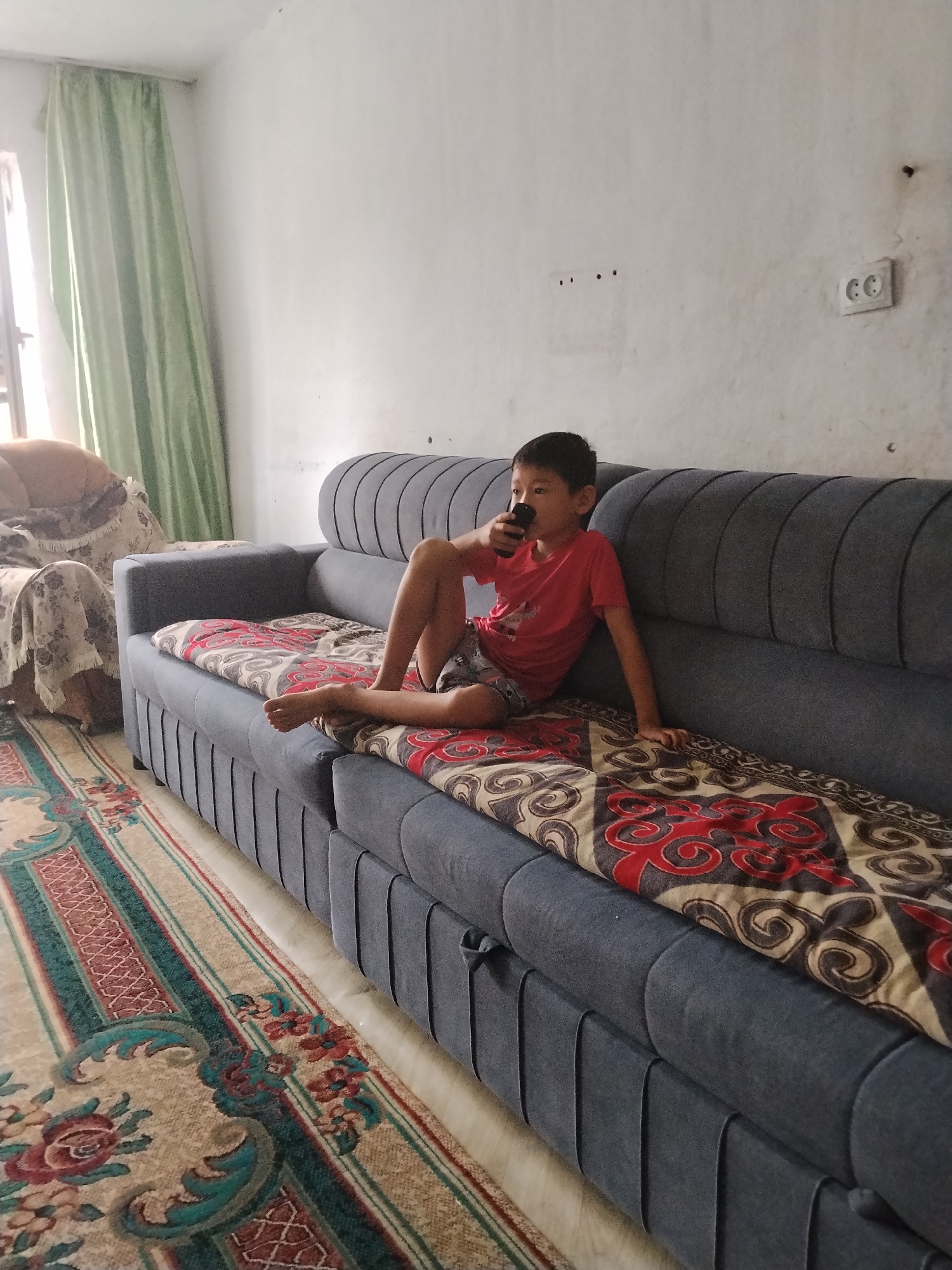 A young boy sits on a dark gray sofa, holding a black remote control to his mouth. He is wearing a red t-shirt and gray shorts, with his legs bent and feet bare. The sofa is covered with a patterned throw featuring red and cream swirls. Next to the sofa is a patterned rug with red and green floral designs. In the background, a window is covered by light green curtains, and a wall with electrical outlets is visible. The scene appears to be indoors, likely a living room, with natural light coming from the window. The boy's expression is neutral as he holds the remote.