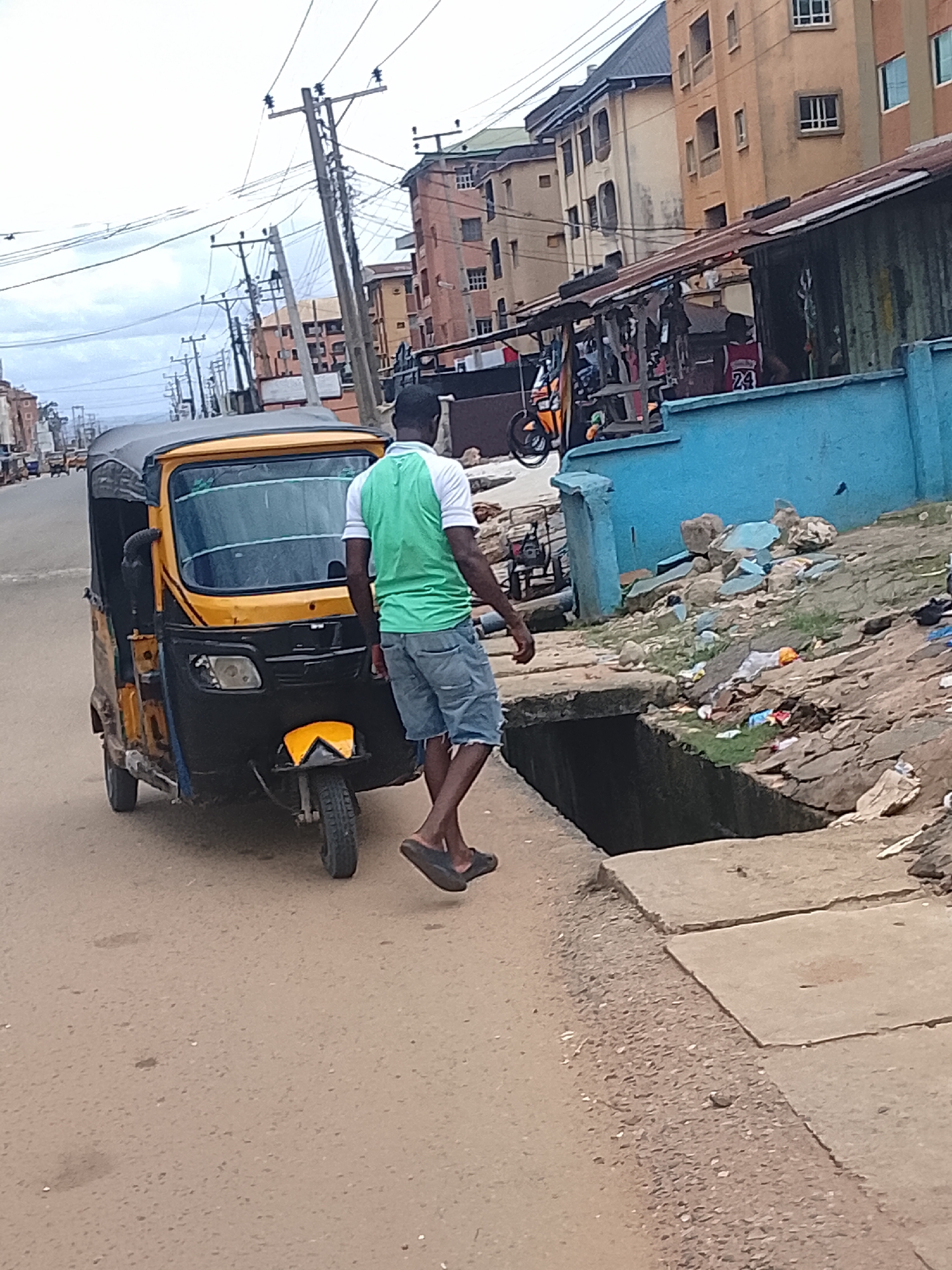 This image depicts a street scene in what appears to be an urban or semi-urban environment, likely in a region with a tropical or subtropical climate, given the overcast sky and the style of buildings.
The primary subjects are a man and a yellow and black auto-rickshaw. The man, seen from behind, is wearing a green and white t-shirt and denim shorts, with dark sandals on his feet. He is walking away from the auto-rickshaw. The auto-rickshaw is parked on a paved road. It has a black canopy, a yellow body, and a black front. There are other vehicles and buildings visible in the background, suggesting a busy street.
The scene appears to be taking place during the daytime, with the sky overcast, indicating a cloudy or possibly rainy weather condition. The surrounding environment shows a mix of residential and commercial structures, with some signs of disrepair, such as the tin roof of a stall and a poorly maintained drainage area with debris. The presence of numerous utility poles with tangled wires also suggests a developing infrastructure.
There is some visible text on the back of a red jersey worn by a person in the background, showing the number "24". The overall impression is one of everyday life unfolding in a bustling, yet somewhat unkempt, urban setting. No strong emotions are conveyed, but the scene captures a moment of mundane activity.