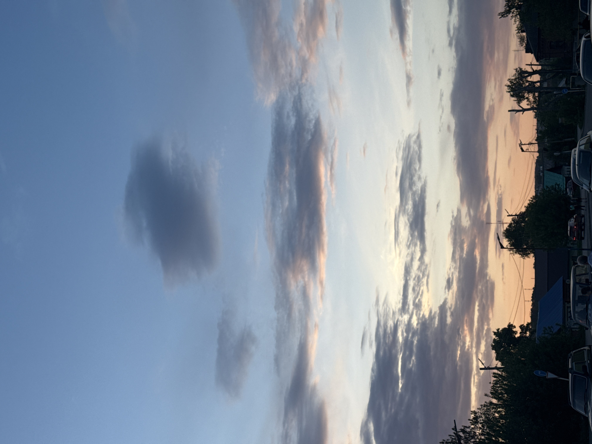 The image captures a scene in Sarapul, Russia. The sky is predominantly blue with varying cloud formations, some tinged with pink hues, suggesting either sunrise or sunset. At the edge of the frame, there is the presence of trees, utility poles, buildings with blue roofs, and parked vehicles.