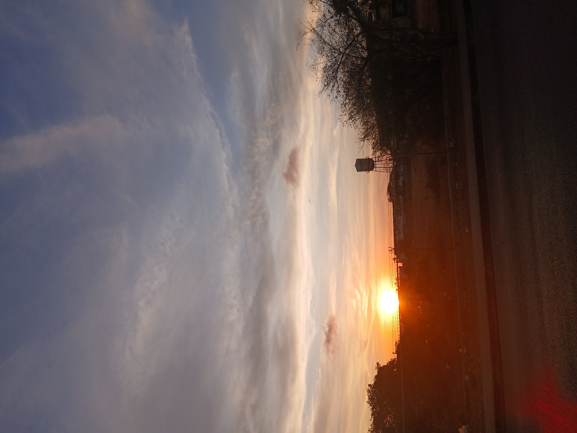 The image depicts a sunset scene, likely in Juan Griego, Venezuela. The sun is setting on the right side of the image, appearing as a bright orange orb obscured slightly by the horizon and trees. The sky is a mix of blue, white, and orange hues, indicative of the sunset. A few bare trees can be seen silhouetted against the sky. A road runs along the right side of the image. A water tower and some buildings are visible in the distance. A fence is separating the road from the horizon.