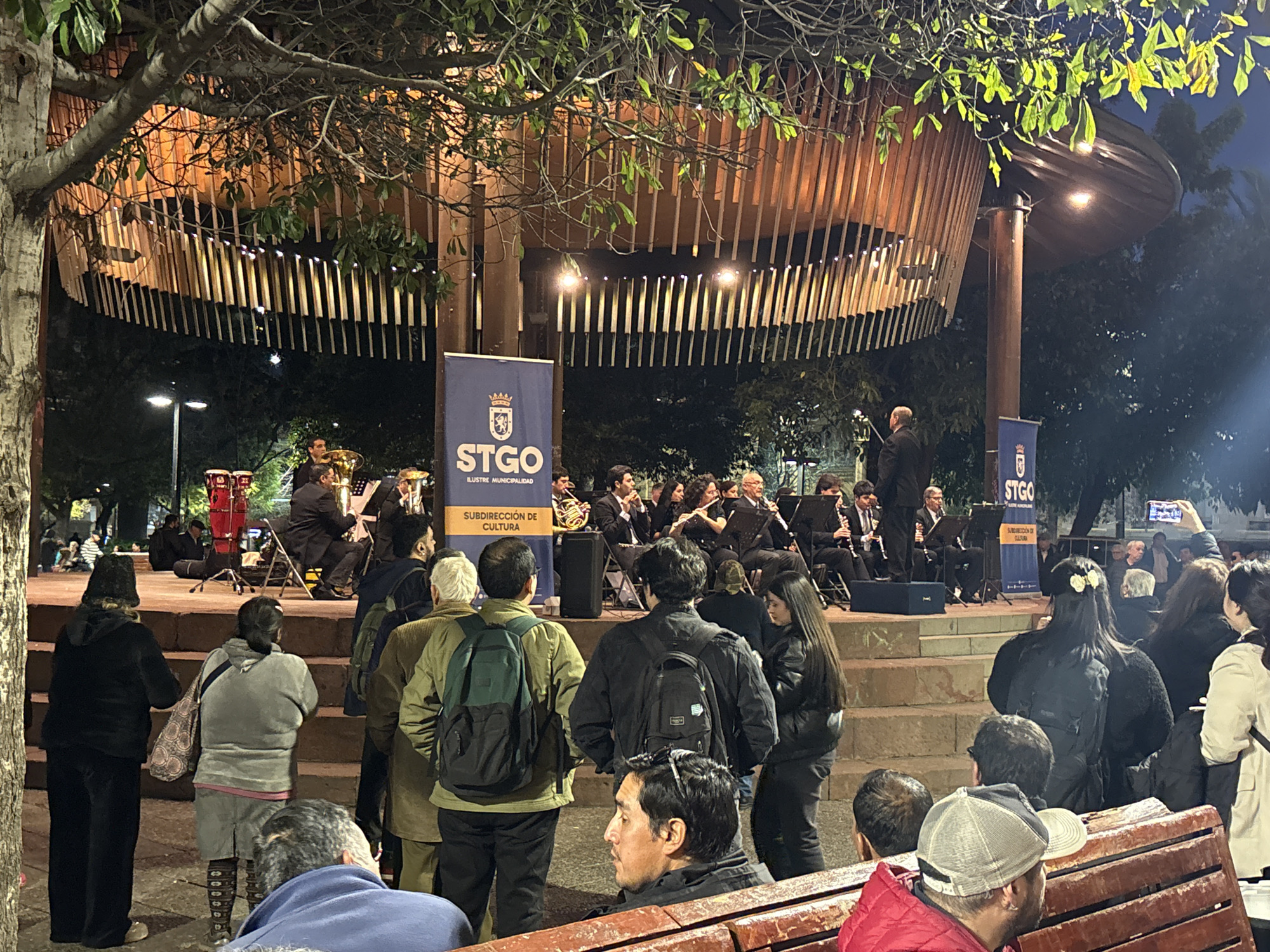 In this evening scene at the Parque Forestal in Santiago, Chile, an orchestra performs on a raised stage in front of a modern, wooden-slatted amphitheater. The lighting casts a warm glow on the performers and the architecture. A blue banner with "STGO" and the text "ILUSTRE MUNICIPALIDAD SUBDIRECCIÓN DE CULTURA" is displayed prominently.
A diverse crowd of approximately 20-30 people is gathered, some standing and others seated on benches in the foreground. Many are observing the orchestra. In the front row, individuals are dressed casually, with one person wearing a red jacket and another a grey baseball cap. To the left, two women are standing, one in black attire with a patterned bag, and another in grey clothing.
The orchestra consists of about 15 musicians playing various instruments, including brass and woodwinds. They are seated on the stage, dressed in formal attire. In the background, trees and streetlights are visible, indicating an outdoor urban setting. The overall atmosphere suggests a community cultural event.