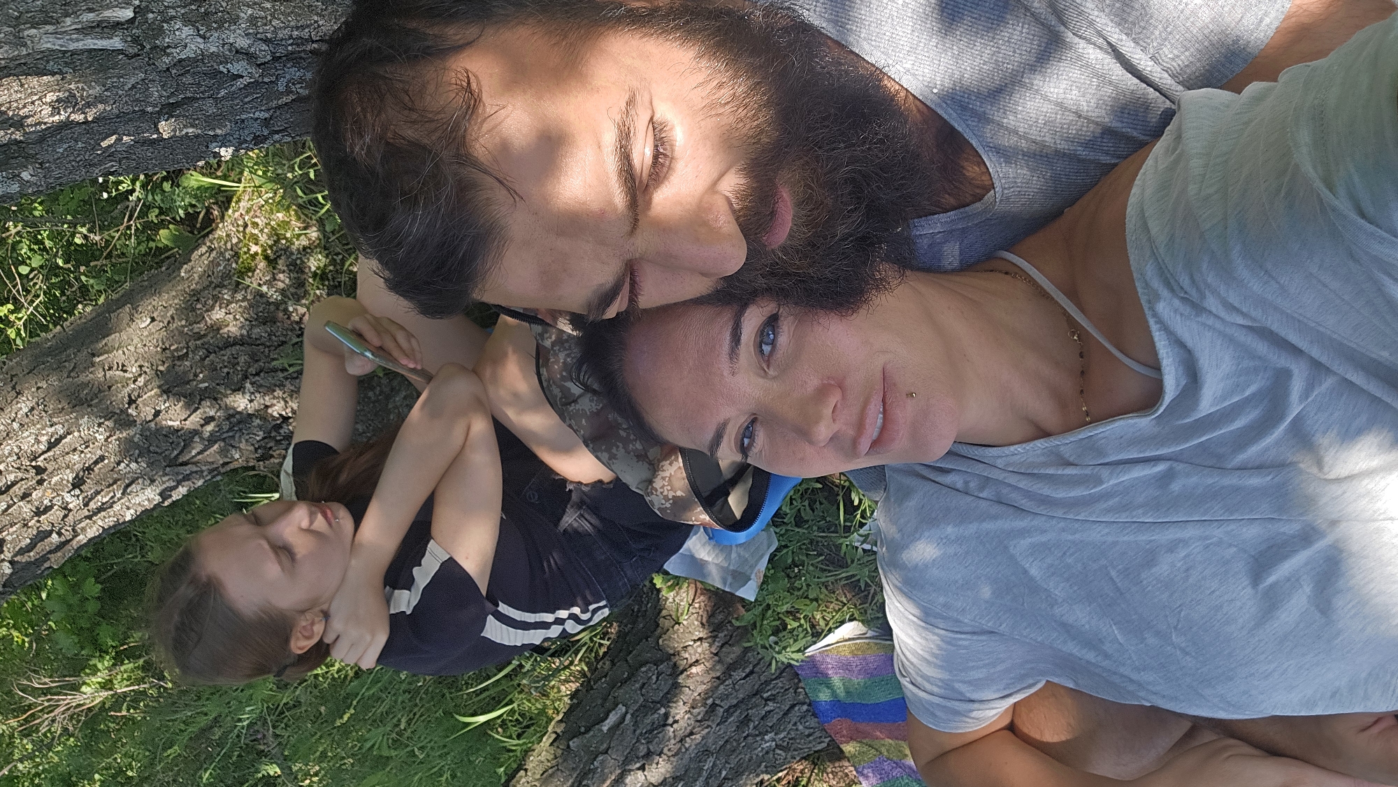In Samara, Russia, three individuals are captured in a close-up, high-angle selfie taken outdoors, likely in a park or natural area. The foreground is dominated by the faces of a man and a woman, positioned in a romantic embrace, their heads tilted towards each other. The man, on the right, has a dark, full beard and is looking down with his eyes partially closed, his face illuminated by dappled sunlight. The woman, in the center, has blue eyes and a lip piercing, and is looking directly at the camera with a subtle smile. She is wearing a light grey t-shirt with a thin gold necklace. Behind them, a younger female is seated with her back to the camera, leaning against a tree. She has long brown hair and is wearing a black t-shirt with white stripes. She is looking away from the camera, holding a smartphone. The environment appears to be a grassy area with large tree trunks visible, suggesting a shaded, outdoor setting. A small, colorful striped blanket and a camouflage bag are partially visible on the ground. The overall impression is one of a relaxed, intimate moment shared amongst family or friends on a sunny day.