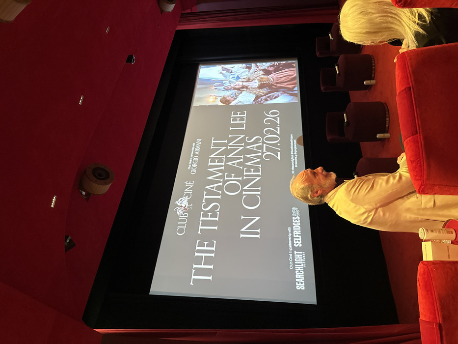 A movie theater shows a promotional screen for “The Testament of Ann Lee” in cinemas on 27.02.26. The movie is sponsored by Club Ciné Giorgio Armani and Searchlight Pictures in partnership with Selfridges&Co. Several rows of plush red seating are partially visible, occupied by an older man in a light jacket and a person with white hair. The theater's red walls feature round, wooden light fixtures.