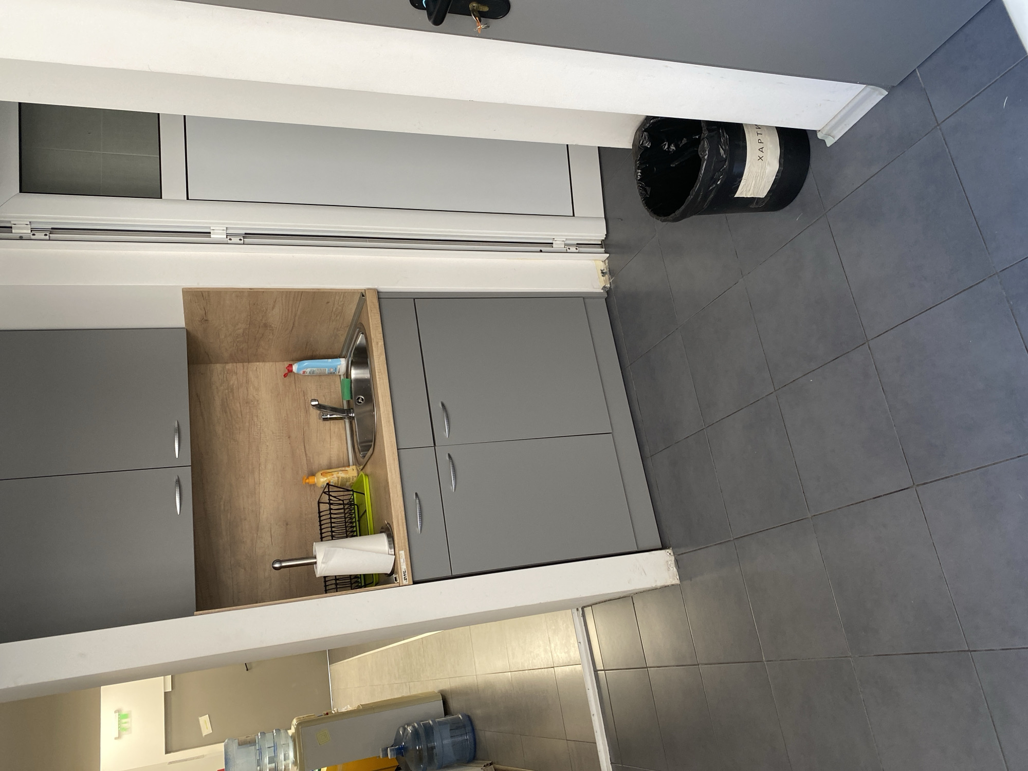The image shows a section of a kitchenette or small kitchen area, oriented with the right side of the view being further away from the camera and slightly higher in the frame than the left side. This is indicated by the perspective of the floor tiles. The color scheme is predominantly grey and white, with some wooden accents.
On the left side, a portion of a kitchen counter and cabinetry is visible. The countertop is light-colored with a wooden-patterned backsplash. A stainless steel sink is present, with a faucet and some cleaning supplies near it. A dish rack containing kitchen items and a paper towel dispenser are also on the counter. Below the counter, there are grey cabinets with elongated silver handles.
Adjacent to the cabinetry, a white door frame is visible. Above this frame is a closed, white door, with a trash bin leaning against the wall on the right side of the door. The bin is black and appears to have a label on it with the word "XAPTH" printed on it.
The floor is covered in large, square, dark grey tiles. A white baseboard runs along the bottom of the cabinets and door frame.
The location is likely Sofia, Bulgaria, as the word “XAPTH” is the Bulgarian transliteration of the word "HARTIYA", which means "paper" in English.